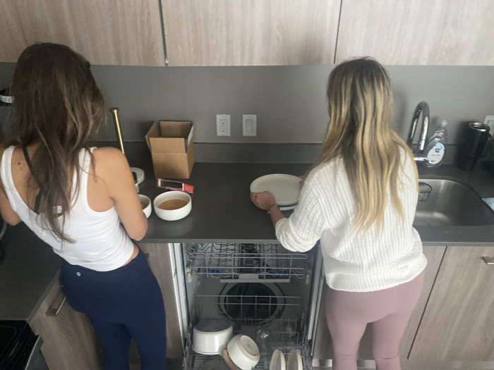 Two women stand in a kitchen. One woman with long brown hair is wearing a white tank top and blue leggings, facing away from the camera. She is near a cardboard box and some small bowls. The other woman, with long blonde hair wearing a white sweater and pink leggings, is putting dishes into an open dishwasher. The kitchen features light-colored wood cabinets, a dark countertop, and a stainless steel sink and faucet.