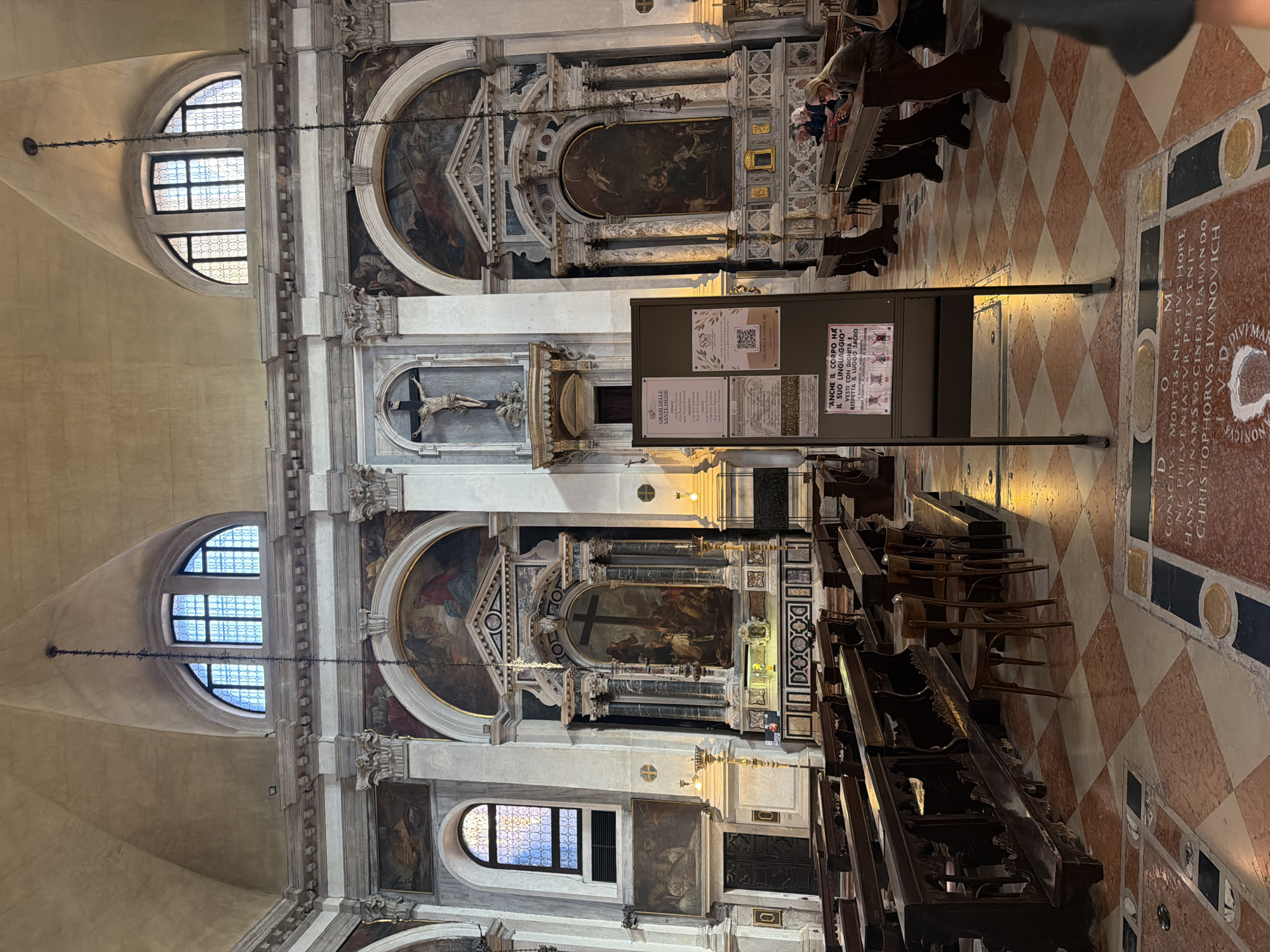 The image shows the interior of a large, ornate church, likely in Venice, Italy, given the checkered marble floor and the architectural style. In the foreground, wooden pews face an altar area. To the left, a large painting is displayed above a dark wooden door. The altar itself is elaborate, featuring a large painting of a crucifixion scene surrounded by columns and decorative elements. Above the altar, a crucifix with a figure of Christ is mounted on the wall. To the right of the altar, another painting is visible. A display board with information, including a QR code, stands prominently in the center of the frame. Several people are seated in the pews in the background, some engaged with their phones. The lighting suggests it is daytime, with light filtering in from arched windows high on the walls. The overall impression is one of a historic and sacred space.