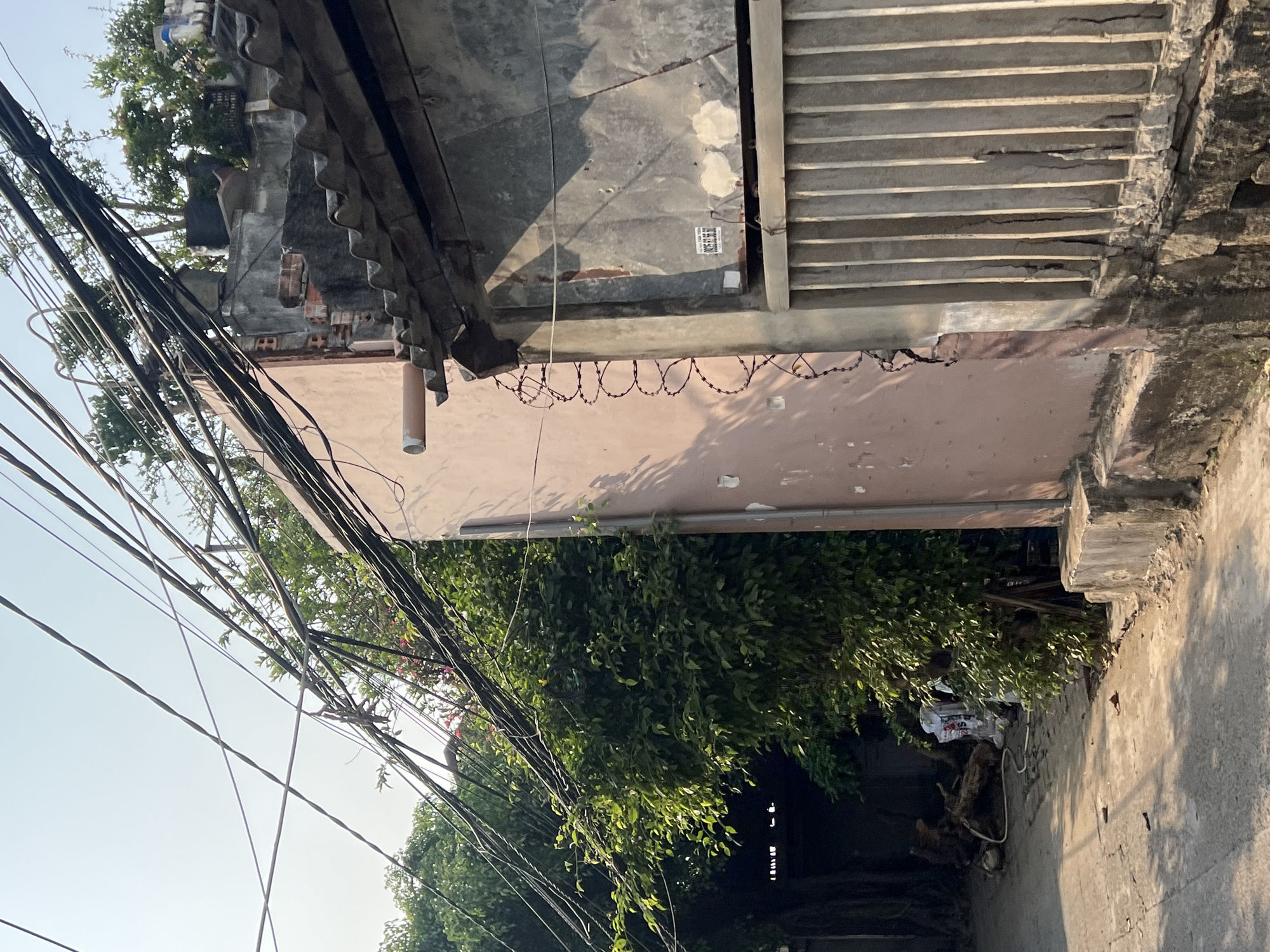 The image shows a street scene in Bắc Nha Trang, Vietnam. Overhead power lines are densely crisscrossed against a light blue sky. A building with a corrugated metal overhang and a pink wall runs along the right side of the frame. Barbed wire is strung along the top of the wall. Green foliage obscures part of the building and the street below. A concrete sidewalk runs alongside the building, leading to a darker area under the foliage, where debris and what appear to be discarded items are visible.