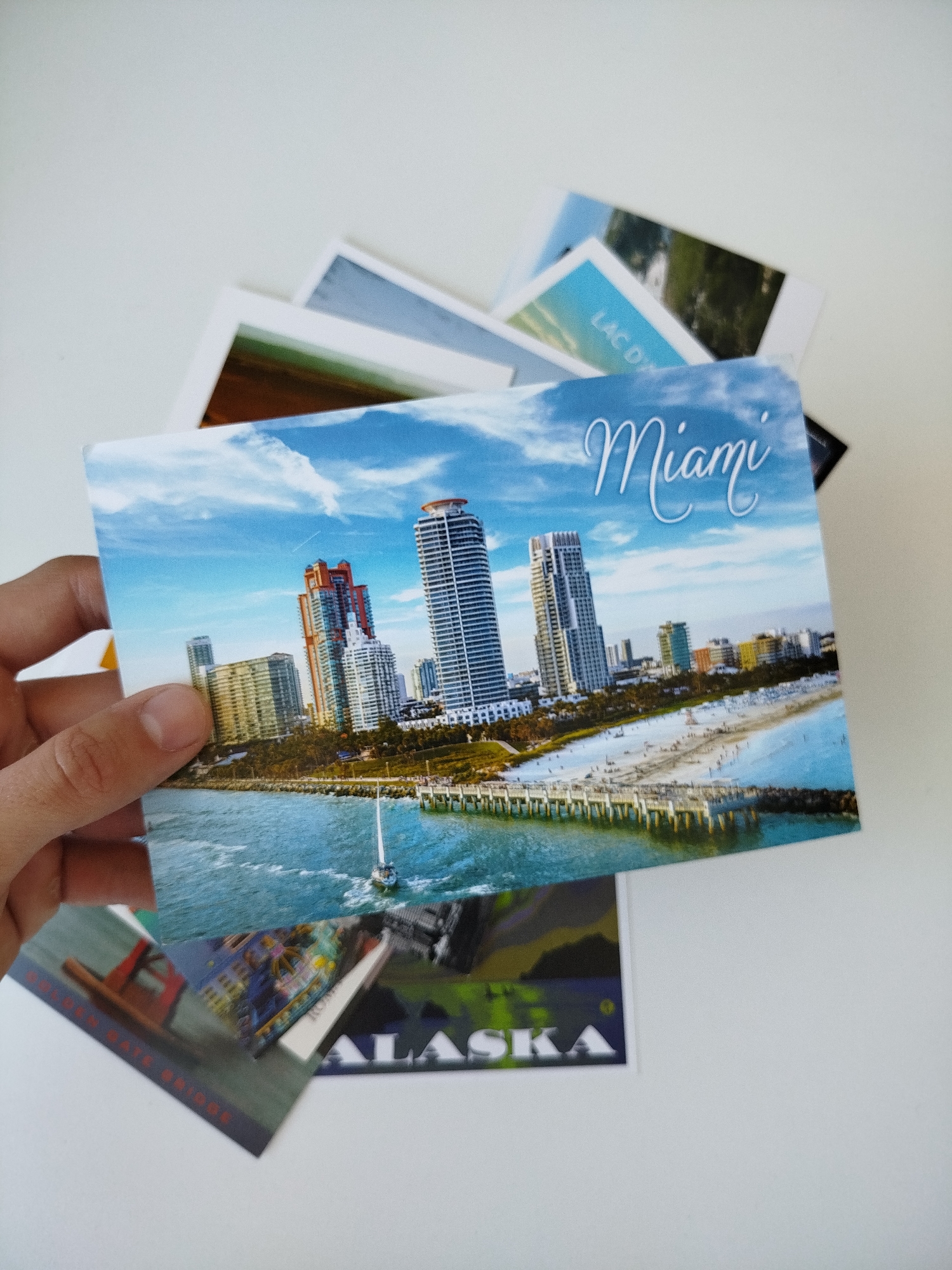 A hand holds a postcard depicting the city of Miami, Florida, USA. The postcard features a cityscape with high-rise buildings, a beach, and a body of water. Stacked beneath the Miami postcard are others, with text visible on one reading "ALASKA". Another partially visible postcard shows an image of what appears to be the Golden Gate Bridge. Other postcards are also partially visible. The background is a plain white surface. This scene is not located in Varna, Bulgaria.