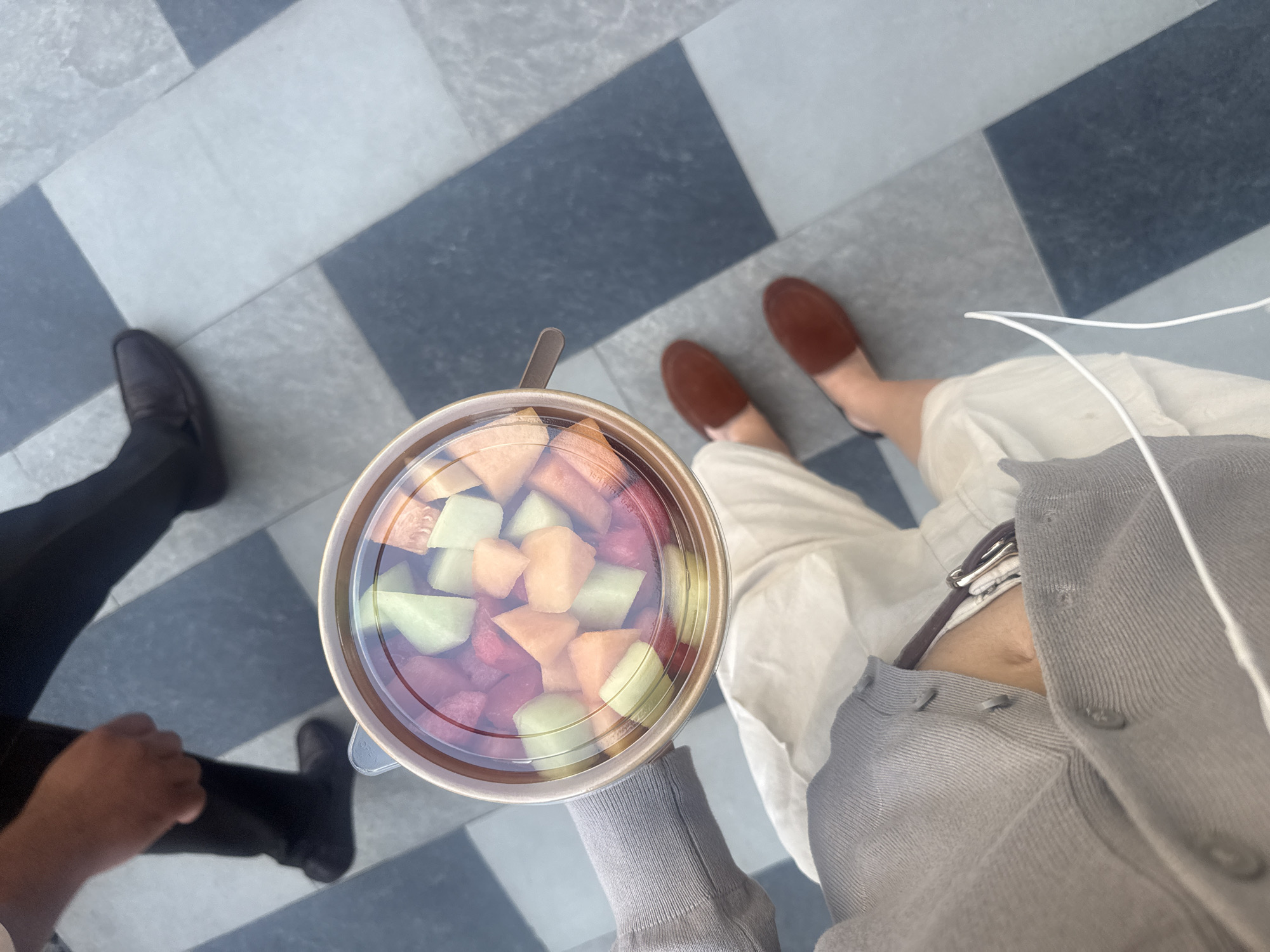 The image, taken from a first-person perspective, features a hand holding a clear plastic container of mixed fruit, including watermelon, honeydew, and cantaloupe. The fruit salad appears fresh, with a slightly glossy surface due to the juice. The container sits against the backdrop of a floor made of square tiles in alternating shades of gray and dark gray. To the left of the fruit container, a person with dark-colored shoes and black pants is partially visible. To the right, another person is visible from the knees down, wearing light-colored trousers and brown slip-on shoes. A portion of a gray cardigan and a white cord, likely from earbuds or headphones, are visible on the right. The location is potentially in the Business Bay area of the United Arab Emirates, given the modern tile work which is commonly found in modern offices and building.