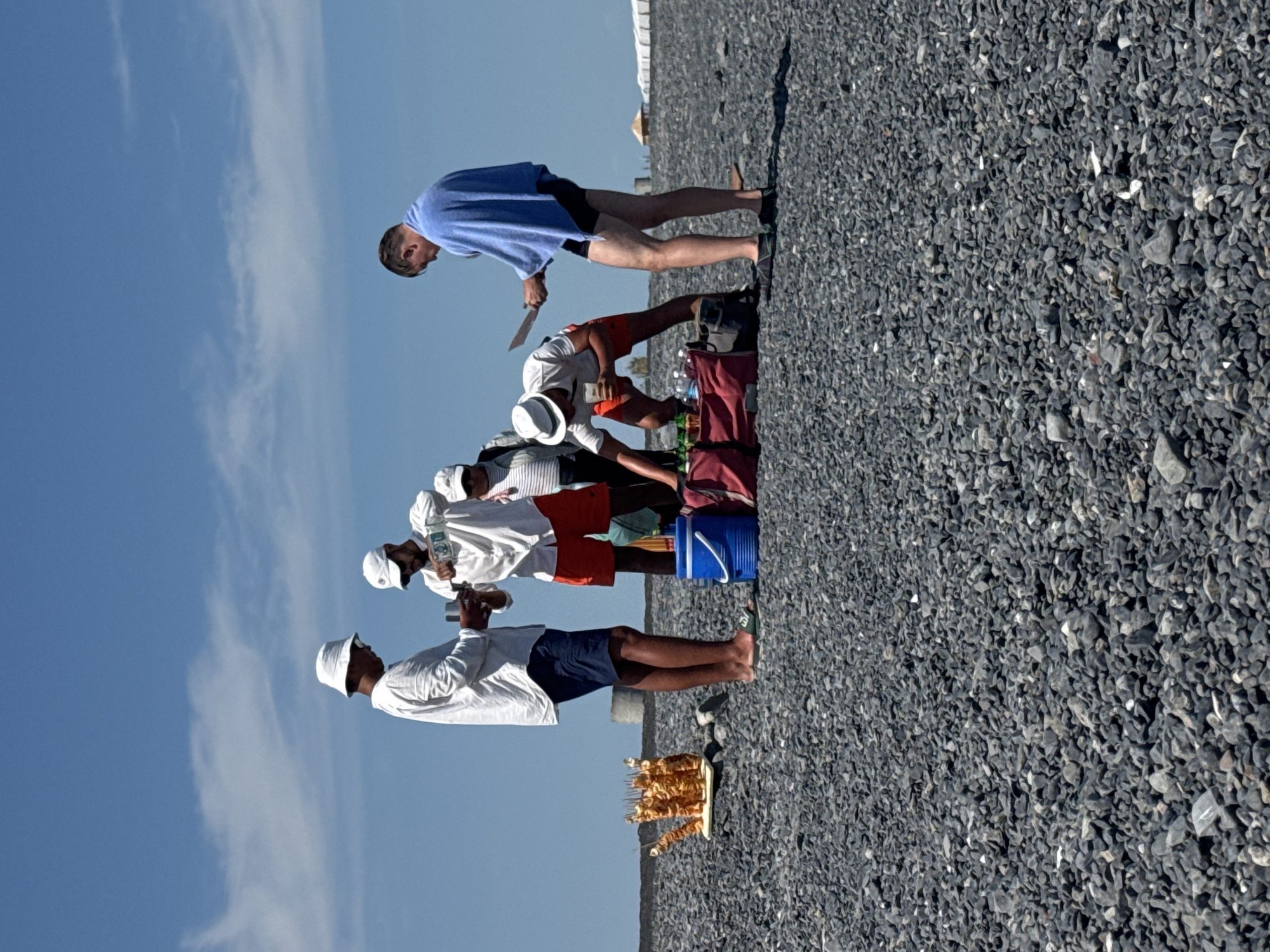 Four men are gathered on a rocky beach under a clear blue sky with scattered clouds. One man on the far left, wearing a white shirt and dark shorts, holds a glass. Next to him, another man in a white shirt and orange shorts is pouring liquid from a bottle into a glass. A third man in a white cap and striped shirt stands behind them, and a fourth man, wearing dark shorts and a blue towel draped over his shoulders, holds a knife.
In front of the men is a collection of items on the ground: two large red bags, a blue cooler, several bottles, and a tray of what appears to be fried potato spirals on skewers. The ground is covered in dark gray pebbles. In the background, a few low structures and white tents are visible in the distance. The scene appears to be a casual gathering or picnic on the beach. The location is identified as a beach in the city of Ақши, Kazakhstan.