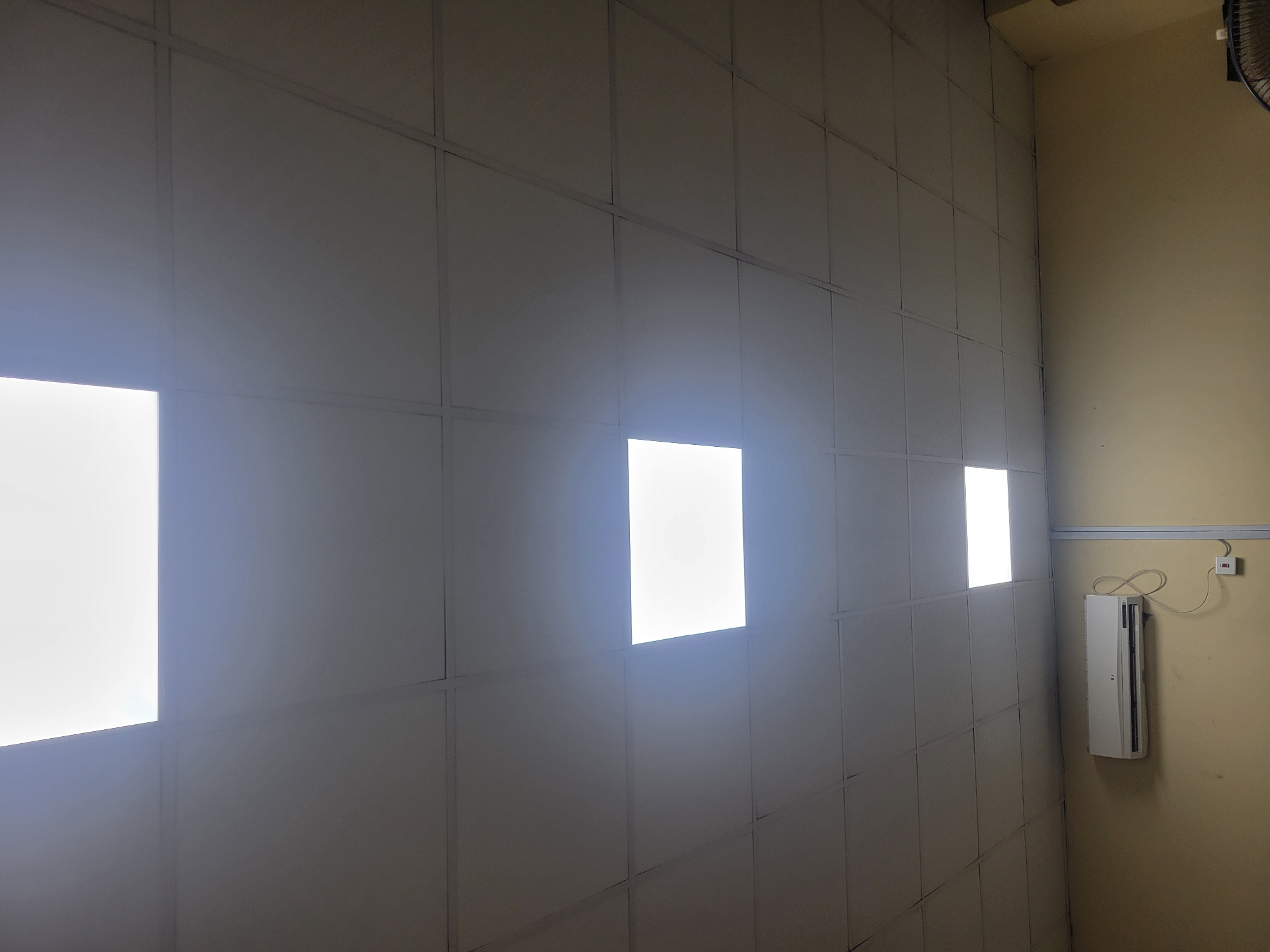 The image depicts a room with a tiled wall that has three illuminated panels. A white air conditioning unit is mounted on a light yellow wall in the corner of the room, to the right of the tiled wall. A red and white light switch is also visible on the yellow wall above the air conditioner. The lighting suggests it is daytime, but the indoor setting makes it difficult to ascertain specific time or weather conditions. The scene appears to be a functional space, possibly an office or a utility area, given the presence of the air conditioner and the utilitarian design of the room. There are no people visible in the image, nor any discernible text. The overall mood is neutral and functional.