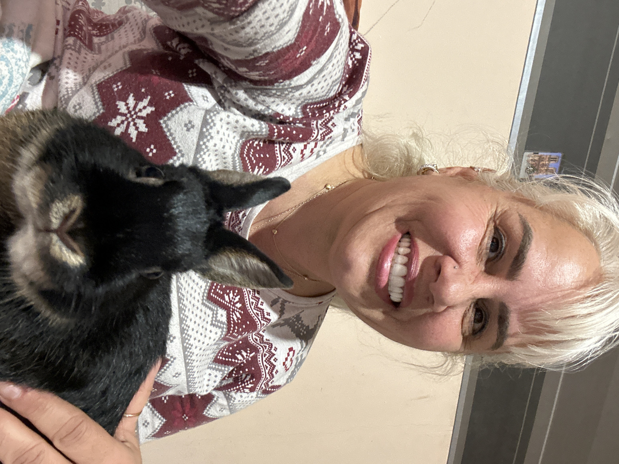 A woman with blonde hair is holding a rabbit. The woman is wearing a long-sleeved shirt with a red and white patterned design. The rabbit is dark brown and black. The background is a neutral beige wall. The photo appears to have been taken indoors with natural light. There is no indication of the location being in Sofia, Bulgaria.