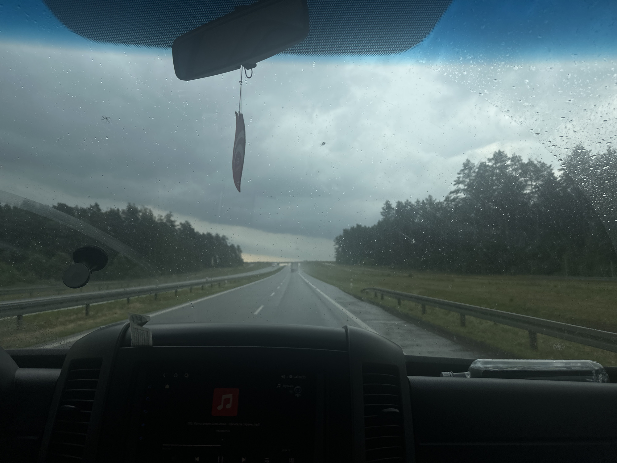 The image is taken from inside a vehicle, looking out through the rain-streaked windshield. The view is of a highway with trees lining both sides. The sky is overcast, suggesting a cloudy or rainy day. In the foreground, the dashboard of the vehicle is visible, with a dark screen displaying what appears to be a music player interface. A car air freshener hangs from the rearview mirror, and a suction cup mount is attached to the windshield. A glass bottle lies on the dashboard. The overall impression is a journey on a highway in potentially inclement weather. The location is Jarnuty, Poland.