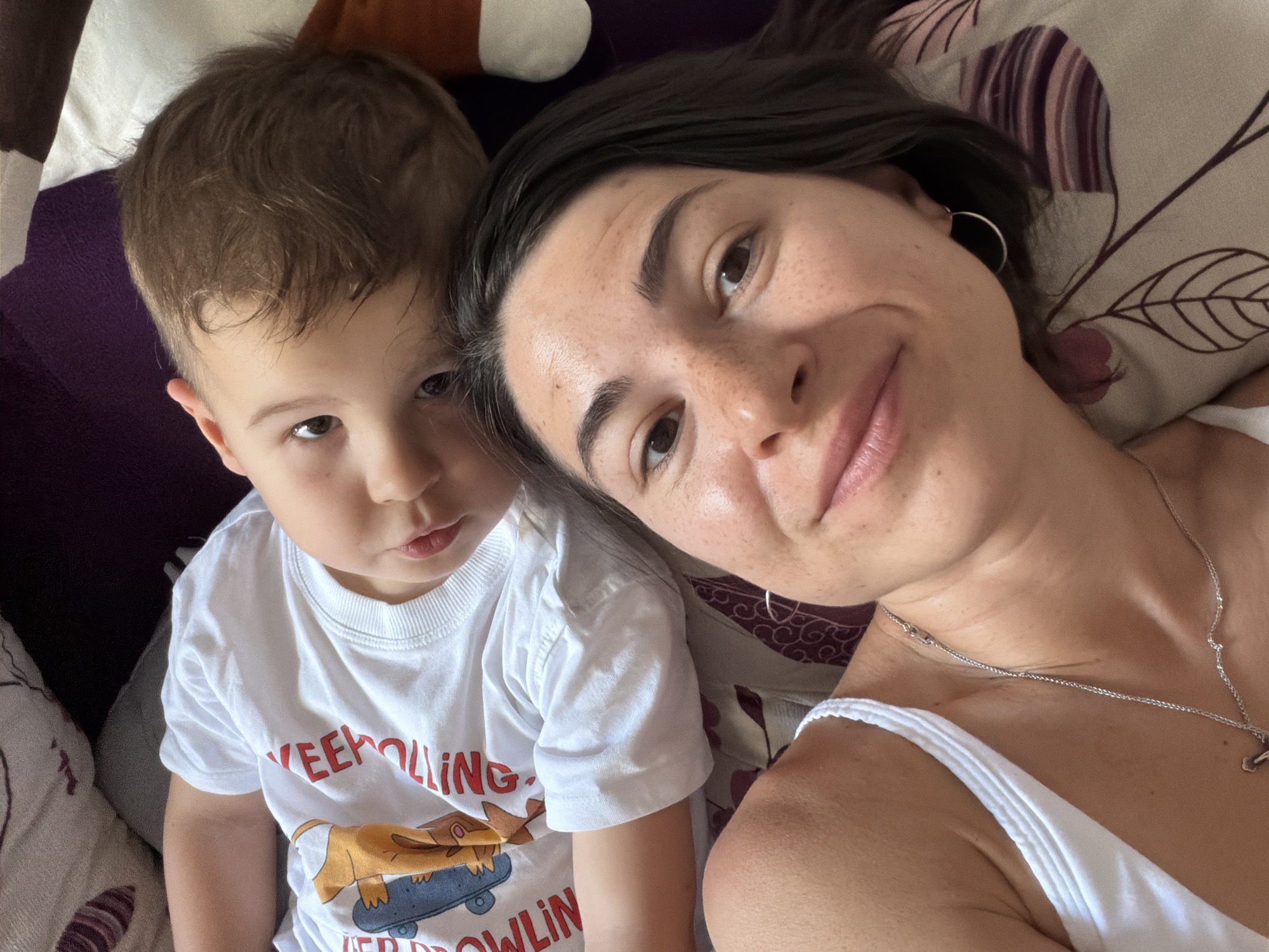 A selfie shows a woman and a young child indoors, likely in a domestic setting in Budva, Montenegro. The woman, positioned on the right side of the frame, has dark hair, fair skin with freckles, and is wearing a silver hoop earring and a necklace with a small charm. She has a slight smile. The child, on the left, has brown hair and is wearing a white t-shirt with a cartoon illustration and the words "KEEPOLLING / EERDROLLIN". Both are looking towards the camera. They appear to be lying on a bed with patterned pillows or bedding, including leaf designs in purple and beige. Other pillows or furniture in shades of brown and purple are visible in the background.