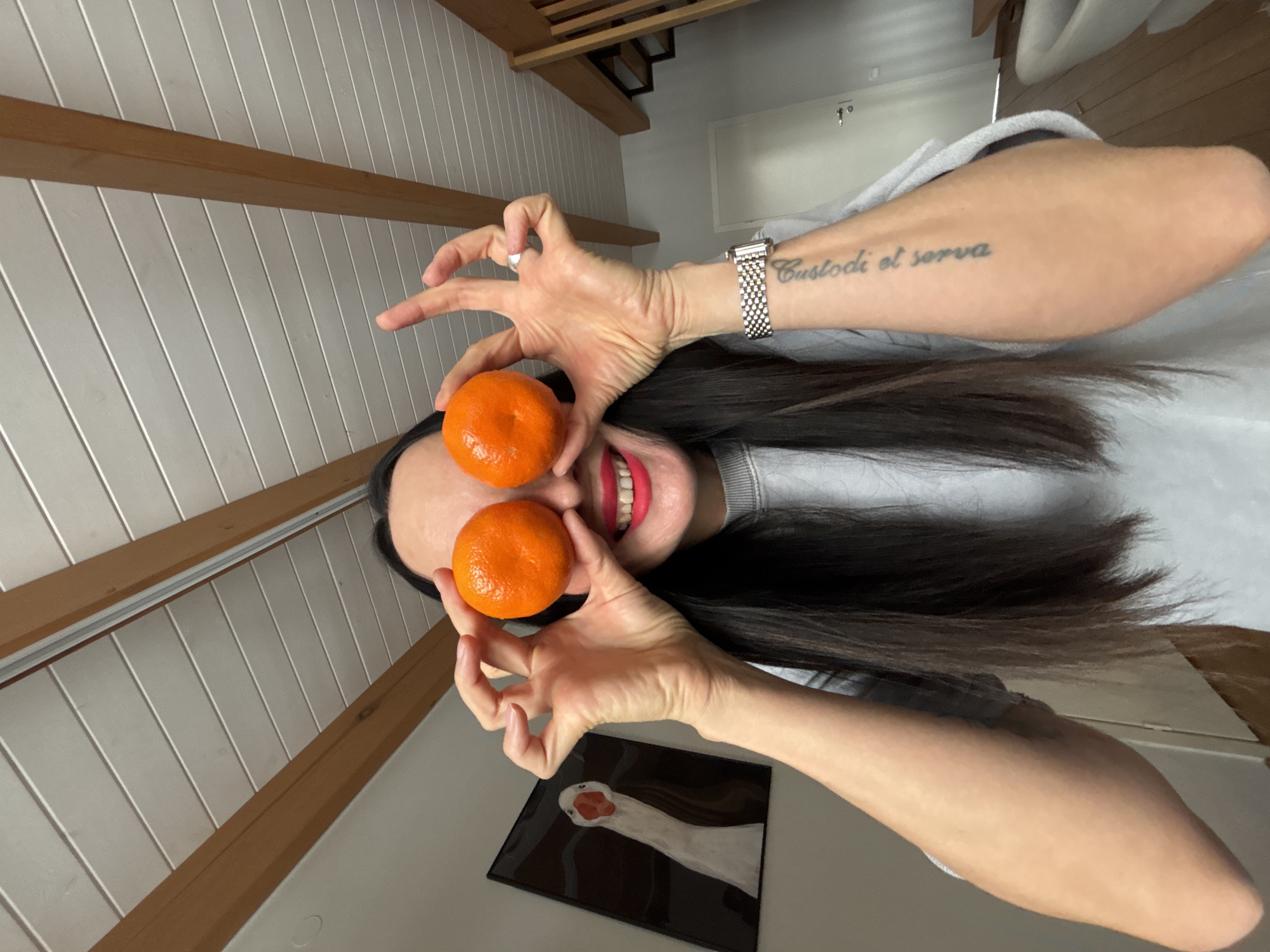 A person is holding two tangerines in front of their eyes. The person has dark hair and is wearing a grey sweater. A watch and a tattoo reading "Custodi et serva" are visible on their left arm. The background includes a white wooden wall and a wooden beam. A framed picture with a white and red design is on the table. This is likely inside a home, but there is no indication of Chaikovsky, Russia.