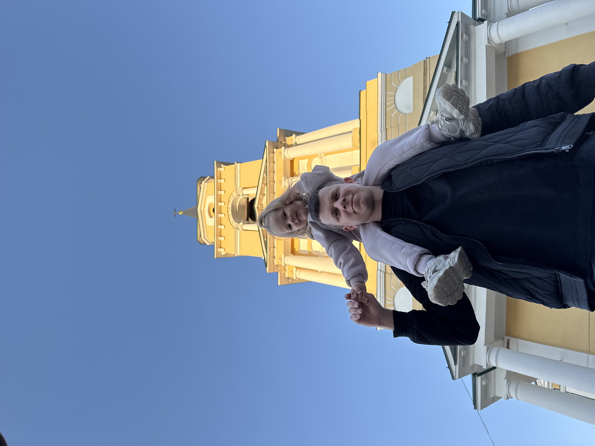 A father carries his young daughter on his shoulders in front of a light yellow building with white columns under a clear blue sky. The building is the Transfiguration Cathedral in the city of Perm, Russia. The father wears a black vest and long-sleeved shirt. The daughter wears a light pink sweatshirt and light gray sneakers. They hold hands. The photo is taken from a low angle, looking up at the subjects.