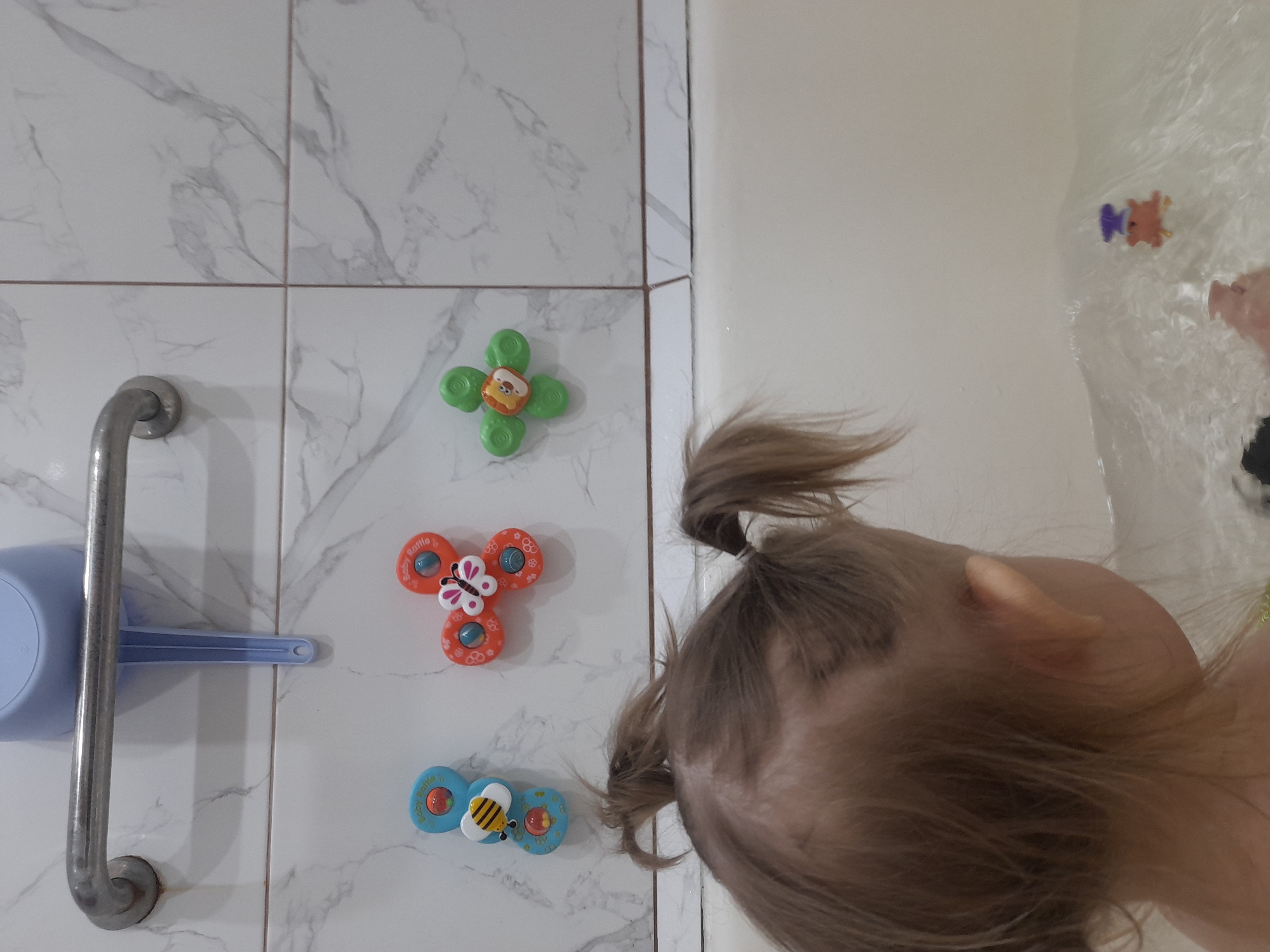 A photograph taken indoors shows a child with light brown hair partially in the frame, positioned to the right of a wall covered with white marble-patterned tiles. On the wall to the left are several bath toys with suction cups, including a green four-leaf clover shape with a lion face, an orange butterfly shape, and a blue bee shape. Also visible on the wall is a silver grab bar and a blue plastic scoop. The location and city are unknown.