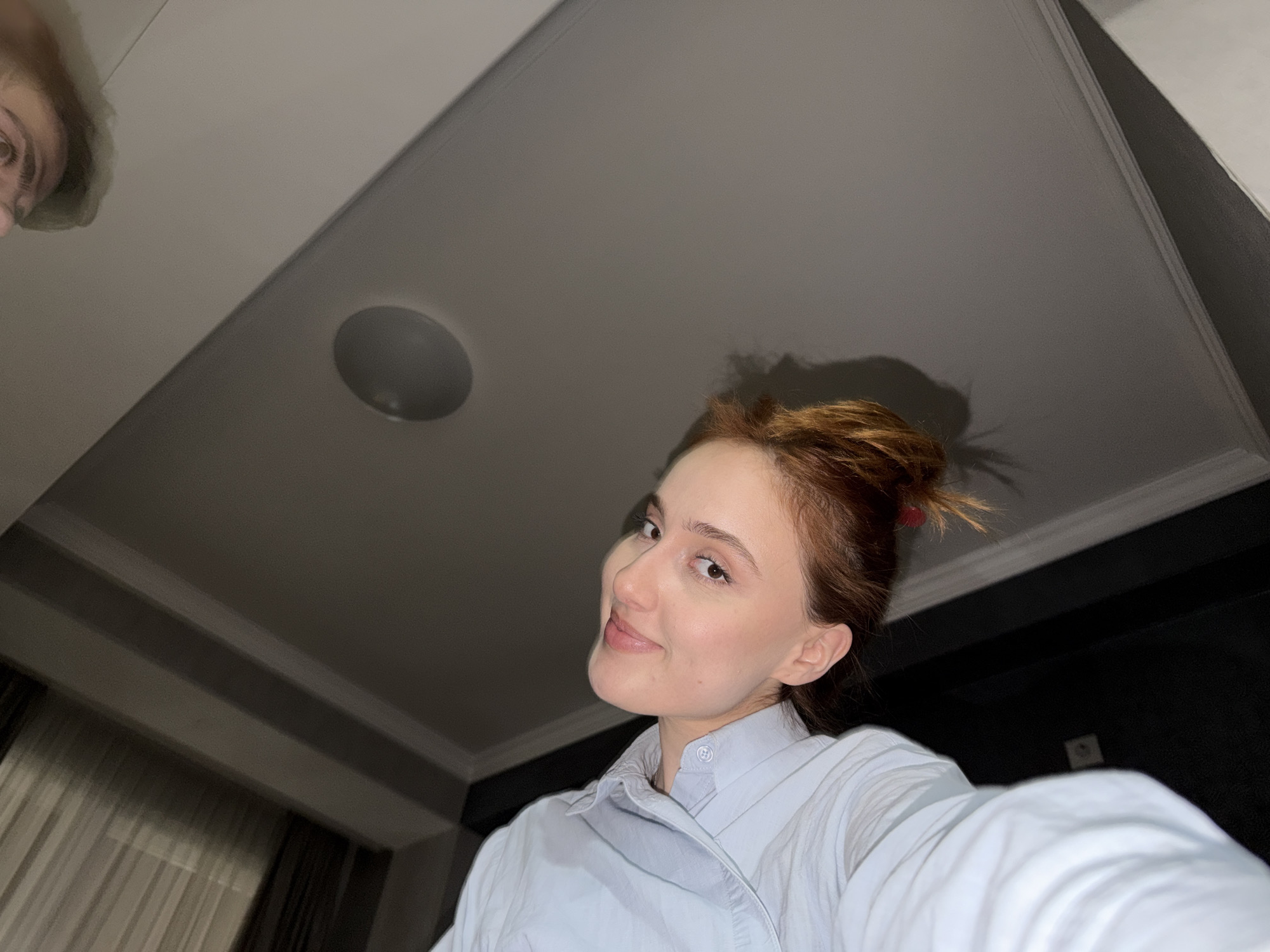 A young, fair-skinned woman with auburn hair pulled back into a messy bun smiles at the camera. She wears a light blue collared shirt and appears to be indoors, likely in a residential setting. The camera angle is from below, looking up at her, capturing the ceiling, a light fixture, and a glimpse of another person's face in the top left corner. Black wall in the background.
No information is present to verify that this image was taken in the city of Tbilisi, Georgia.