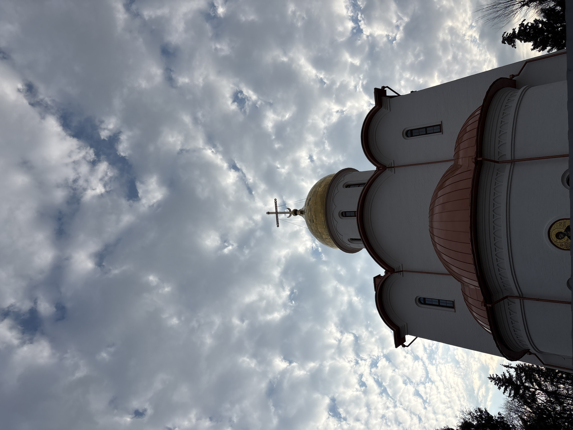 The image depicts a low-angle view of an Orthodox church against a cloudy sky. The church has white walls, copper-colored domes, and a golden central dome topped with a cross. The architectural style suggests it may be located in Eastern Europe, possibly Russia. The sky is filled with numerous small, puffy clouds, and patches of blue are visible between them. The branches of trees are visible in the lower right and upper right corners, partially obscuring the structure. Based on visual cues, the location is likely Ulyanovsk, Russia, as it appears to be consistent with architectural details of buildings found in that city.
