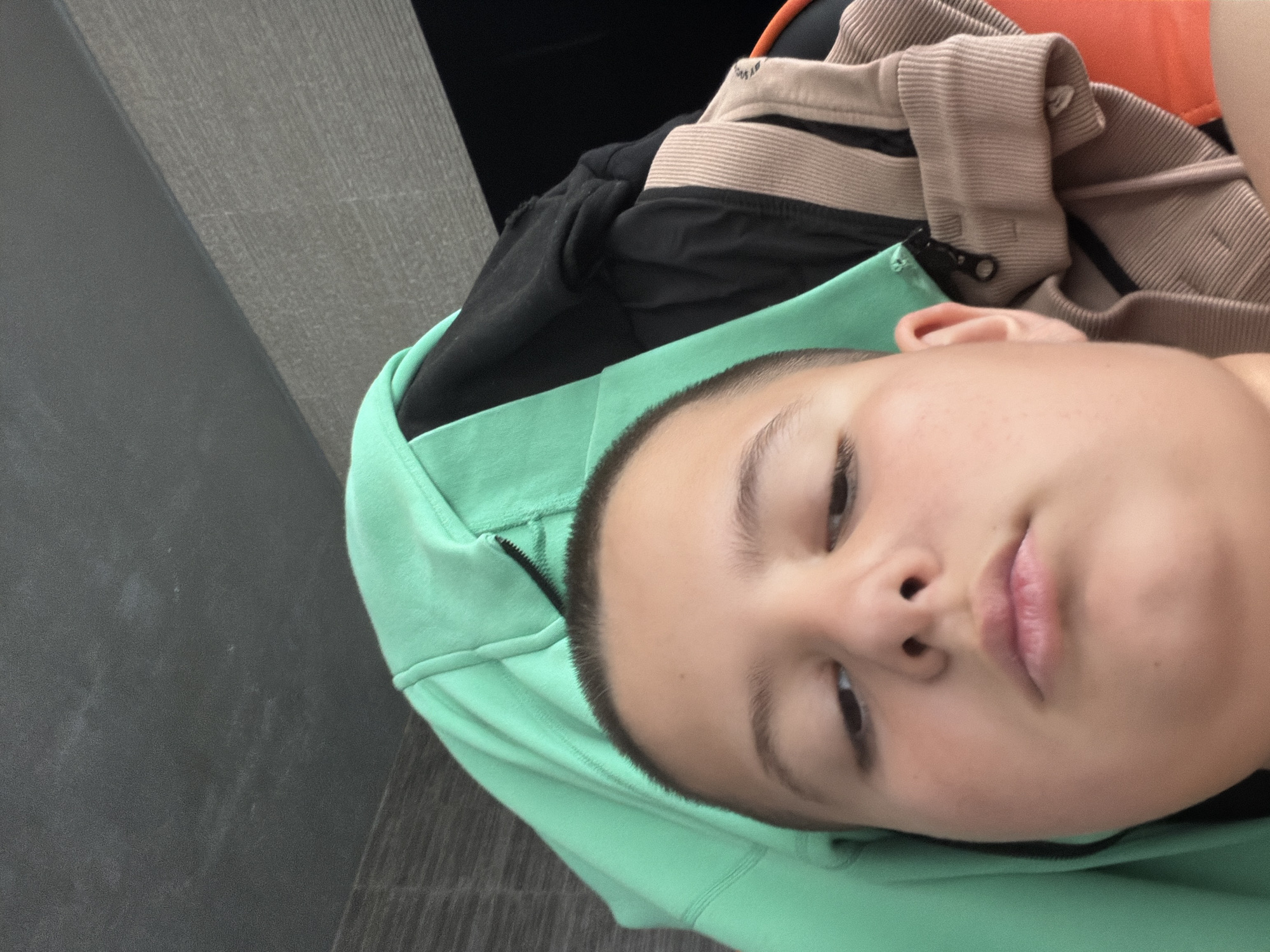 A close-up, angled shot of a person with short, dark hair and light skin. They are wearing a light green hoodie, and other clothes are visible in the background, including items with black, beige, and orange colors. The background includes a gray surface with subtle horizontal lines, and an area of darkness at the top of the frame. The subject is looking slightly downwards towards the camera with a relaxed facial expression. The location is unknown; nothing in the image indicates that it is Shumen, Bulgaria.