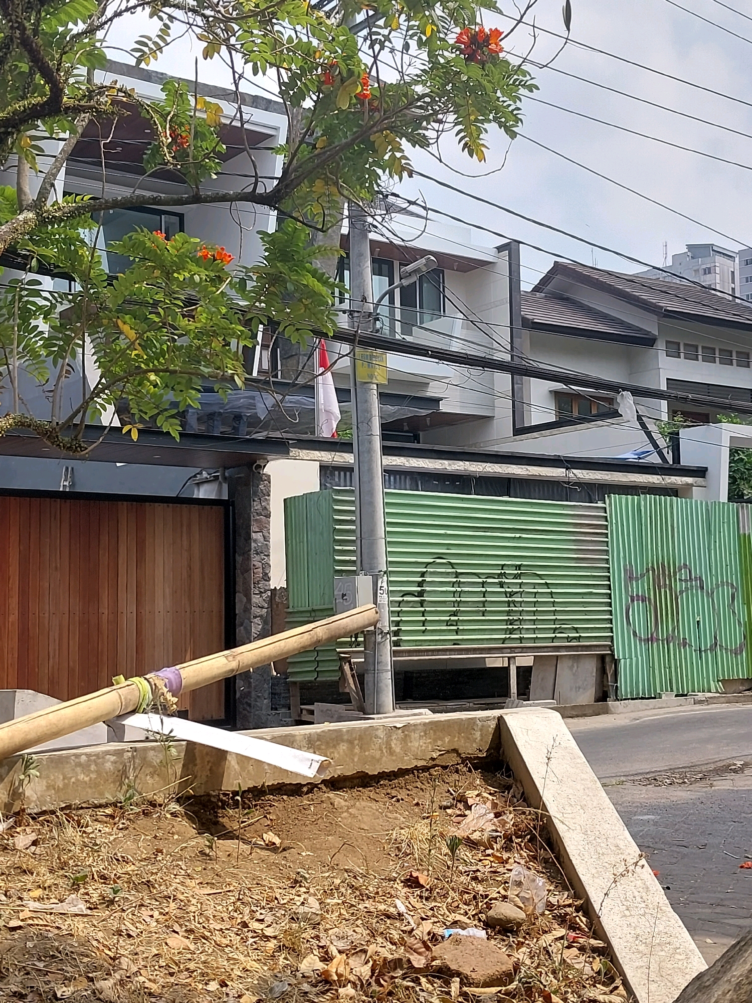 A street view in Malang, Indonesia, shows a modern house with a wooden gate and a green corrugated metal fence. A tree with orange flowers hangs over the scene, with power lines crisscrossing the sky. In the foreground, a bamboo pole rests on a concrete barrier, and the ground is covered with dry leaves and some litter.