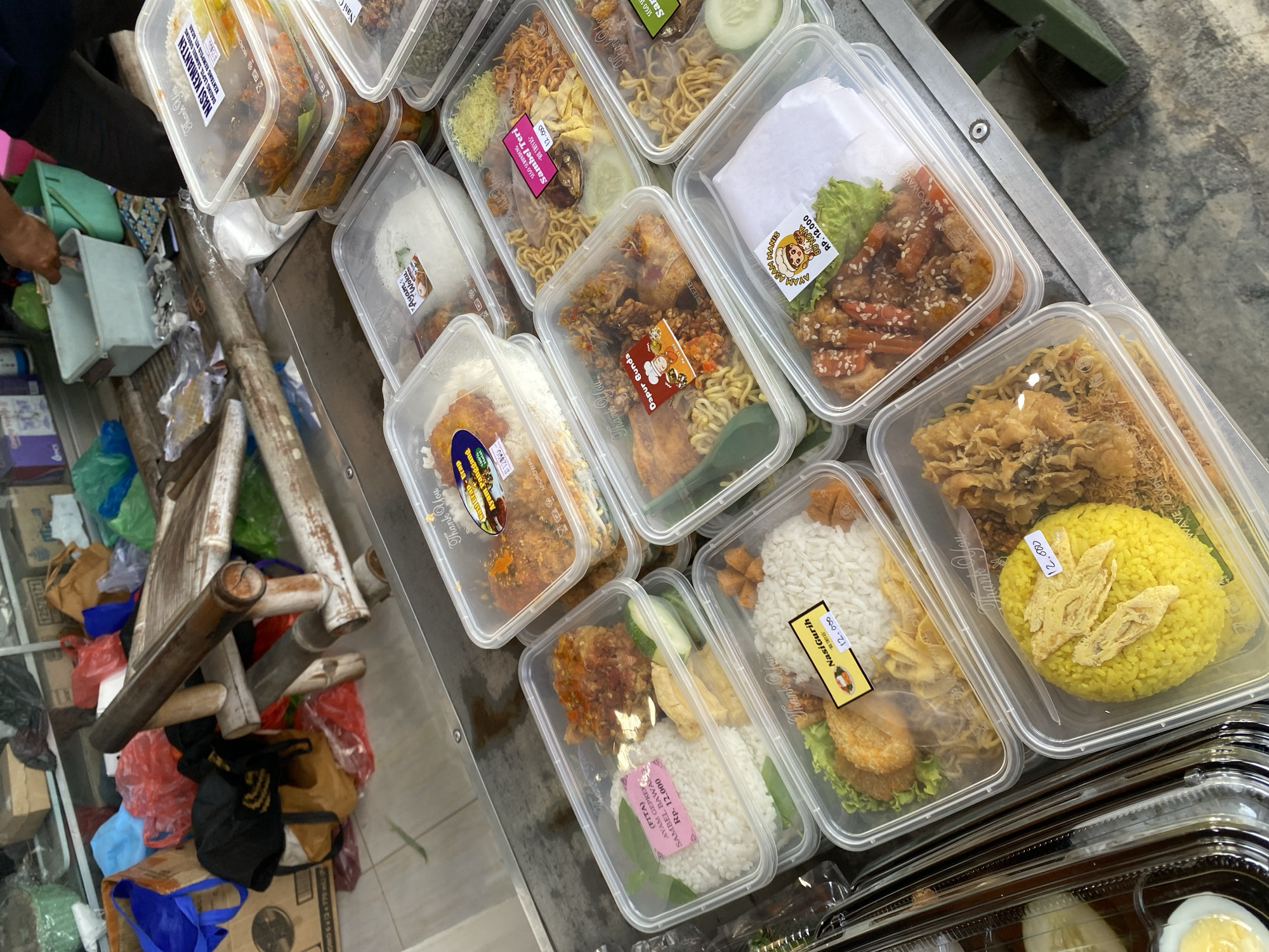 The image displays a selection of pre-packaged meals in clear plastic containers, arranged on a metal table. The meals appear to be of Indonesian cuisine, featuring rice, noodles, and various savory dishes, some of which are fried. Many of the containers have labels indicating the name of the dish and its price, such as "Nasi Kenar," "Ayam Geprek," and "Nasi Gurih." The background suggests an outdoor or semi-outdoor market setting, with blurred figures and shopping bags visible. The overall scene depicts a food stall or market vendor preparing to sell these meals. The location is identified as Calukan RT 1 RW 5 Keboansikep, Indonesia, as suggested by the provided text.