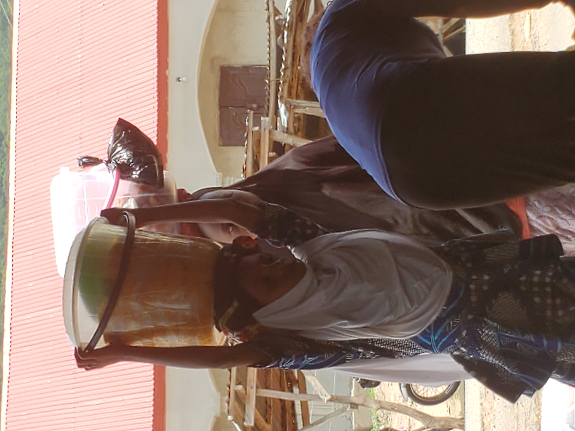 In Mararaba, Nigeria, a woman is carrying a large yellow container on her head. She wears a white headscarf, and a blue and black patterned outfit. Another person wearing a blue shirt and dark pants is partially visible in the frame. A black plastic bag is placed next to the container on the woman’s head. The background includes a building with a red corrugated iron roof and a cream-colored wall. A portion of a motorcycle is visible at the bottom of the image.