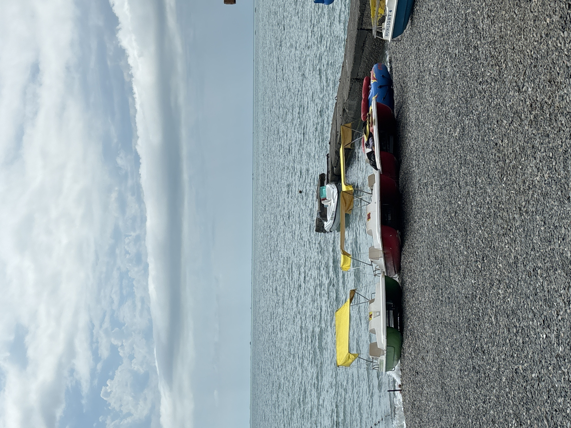 The image depicts a rocky beach in Sochi, Russia, with the Black Sea stretching to the horizon under a cloudy sky. In the foreground, several small pedal boats with yellow canopies are lined up on the shore. One green pedal boat is on the left, followed by two red ones. A blue inflatable tube rests on the beach near the red boats. To the right, a white motorboat with the word "SUNLIGHT 4" visible on its side is partially in view. A concrete pier extends into the water from the right side of the frame, with another small motorboat docked there. The water is calm, and distant boats or structures can be seen on the horizon. The overall scene is peaceful and suggestive of a recreational area on a cloudy day.