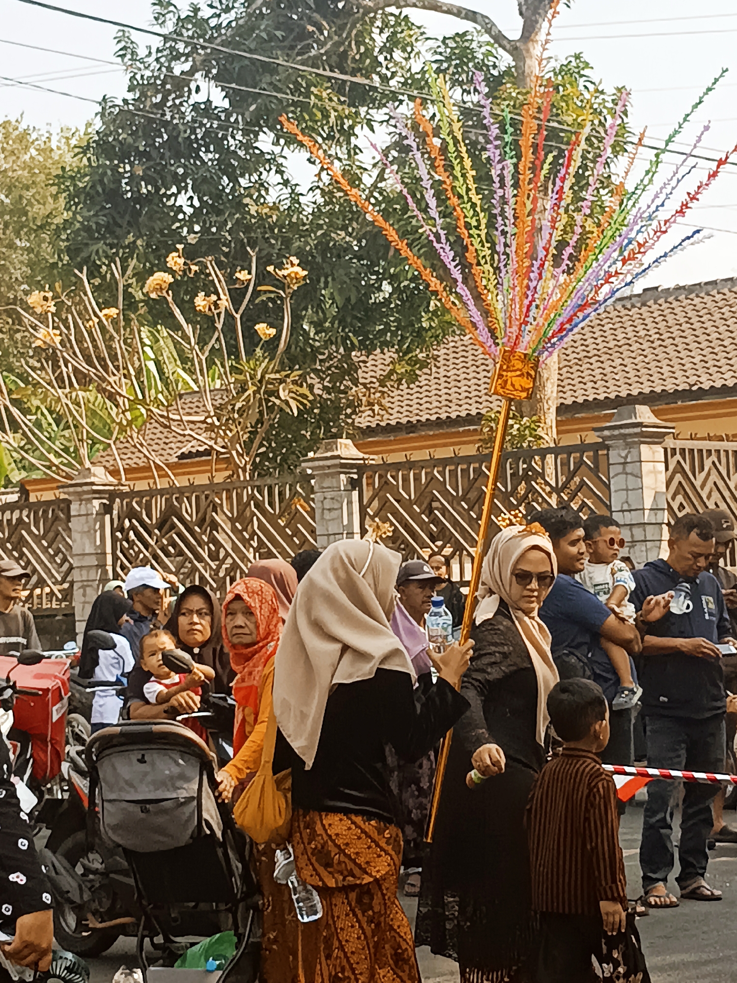 In Jatitengah, Indonesia, a vibrant procession is underway. A large, ornate golden staff adorned with a multitude of colorful paper strips, resembling leaves or feathers, is prominently held aloft, its vibrant display contrasting with the surrounding environment. The crowd appears to be observing or participating in this event. Several individuals are visible, some wearing traditional Indonesian attire, including headscarves and patterned fabrics. A young child is being held, and a baby stroller is present, suggesting a family-oriented gathering. In the background, a fence with an intricate design borders a building with a tiled roof, and lush green trees provide a natural backdrop. The scene is illuminated by daylight, indicating an outdoor event.