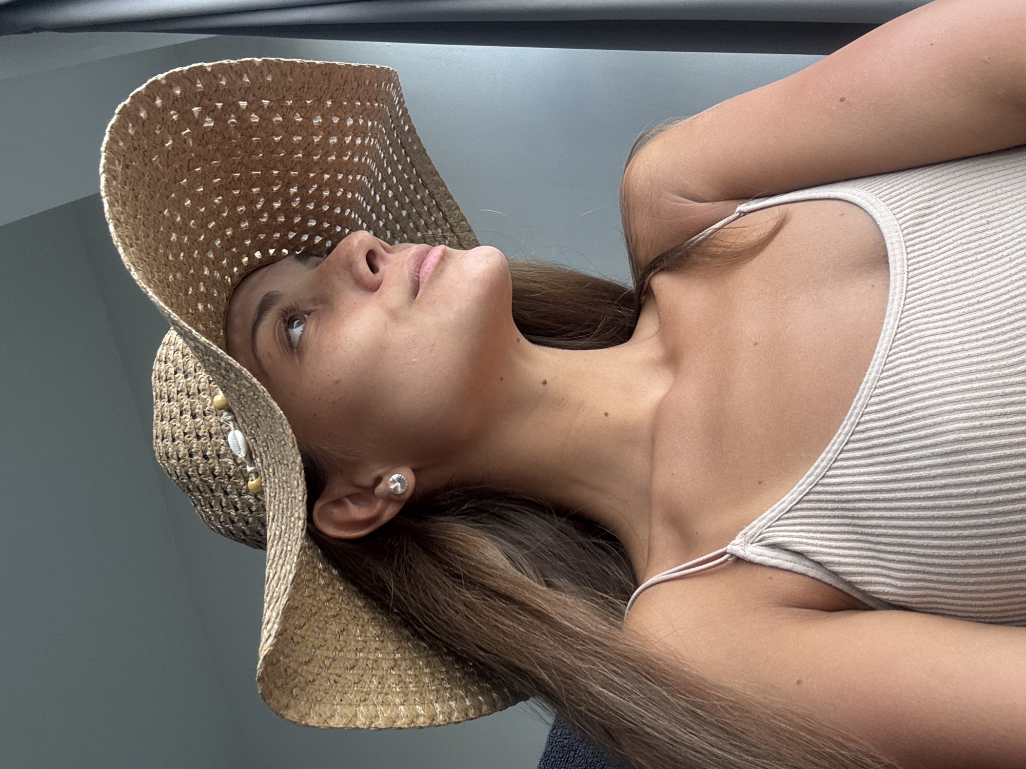 The image is a close-up, angled portrait of a young woman, captured from a low perspective, showing her from the chest upwards. She has long, straight, brown hair, fair skin, and is looking upwards with her head tilted back, her lips slightly parted. She is wearing a light-colored, possibly beige or off-white, horizontally striped tank top with thin shoulder straps. A woven straw hat adorns her head, featuring small, decorative white shells and wooden beads on the side. A silver-colored stud earring is visible in her right ear.
The background is a plain, dark gray or blue surface, suggesting an indoor or sheltered outdoor setting. The lighting is bright and natural, indicating daytime. The woman appears relaxed, seemingly posing for the photograph. There is no visible text in the image. The scene is set in Kuala Lumpur, Malaysia.
