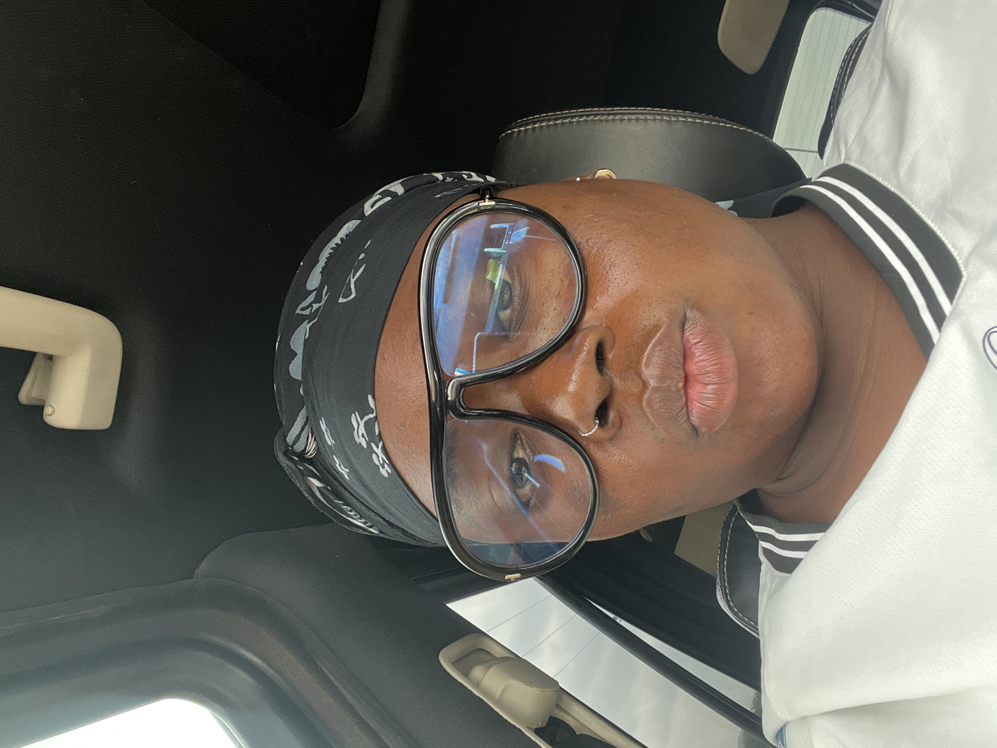 The image presents a close-up portrait of a person with dark skin, oriented sideways within a vehicle, likely a car, in Abuja, Nigeria. The subject wears large, dark-framed glasses with prominent reflections of the sky and surroundings, suggesting it is daytime. A black bandana with a subtle white pattern covers their head. They are dressed in a white garment featuring black and white striped detailing on what appears to be a collar or cuff. A silver nose ring is visible on their right nostril.
The person's expression is neutral, with their gaze directed through the glasses. The immediate background reveals the dark fabric headliner and a light-colored overhead console or grab handle of the car. A dark leather car seat is also partially visible behind the subject. The bright, reflective light on the glasses indicates ample ambient light, consistent with daytime. No discernible text is clearly visible on the clothing or accessories.