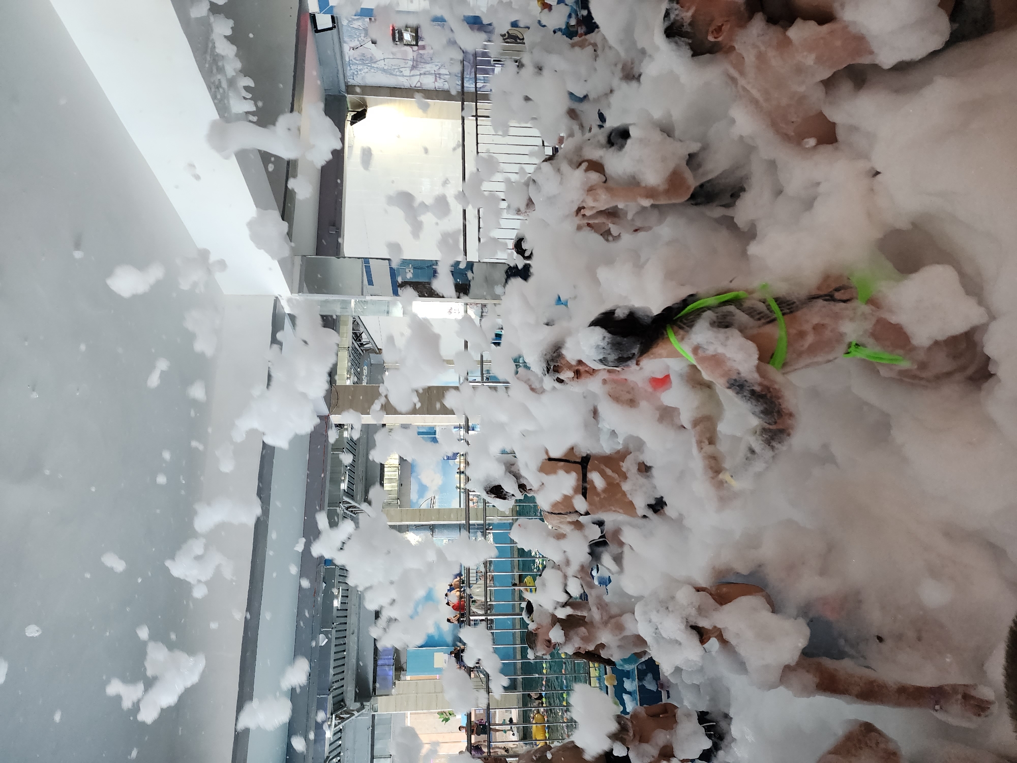 The image captures a foam party in what appears to be an indoor water park, potentially located in Sochi, Russia. Numerous individuals, mostly scantily clad, are immersed in a thick layer of white foam. The setting includes interior architectural elements such as walls, windows, and railings. The foreground and background are dominated by the foam, obscuring finer details. People are interacting and enjoying themselves in the foamy environment.