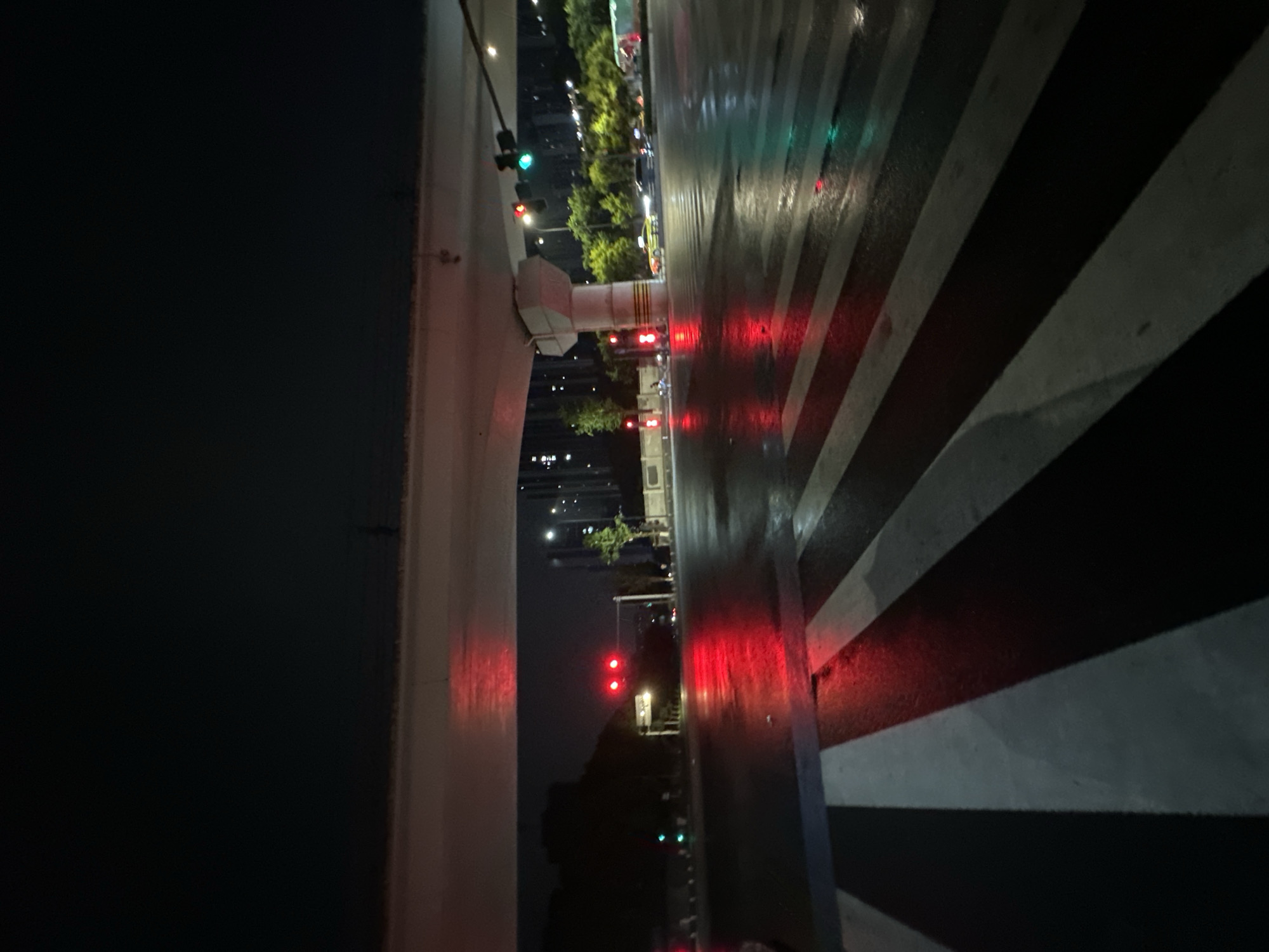 The image captures a nighttime urban scene in the Chongqing High-tech Zone, China. The environment is characterized by a wet, dark asphalt road, indicative of recent rain, causing strong reflections of light sources. In the immediate foreground, a broad zebra crossing with white stripes against the dark road surface is clearly visible.
The midground features a multi-lane road segment situated beneath a large concrete overpass. Two primary red traffic lights are prominently displayed straight ahead, signaling a stop, with their vivid red reflections mirrored on the slick road. To the right, a green traffic light is visible, also casting a reflection onto the wet surface. A substantial, multi-faceted concrete pillar supporting the overpass stands on the right side of the road, featuring yellow and black diagonal warning stripes near its base. Several vehicles, including a distinct yellow-green taxi, are discernible in the mid-distance, either stationary or in motion.
In the background, tall, modern buildings with numerous illuminated windows rise against a dark sky, characteristic of a developed urban area. Scattered urban foliage and streetlights contribute to the ambient illumination. No individuals or specific human actions are discernible within the frame.