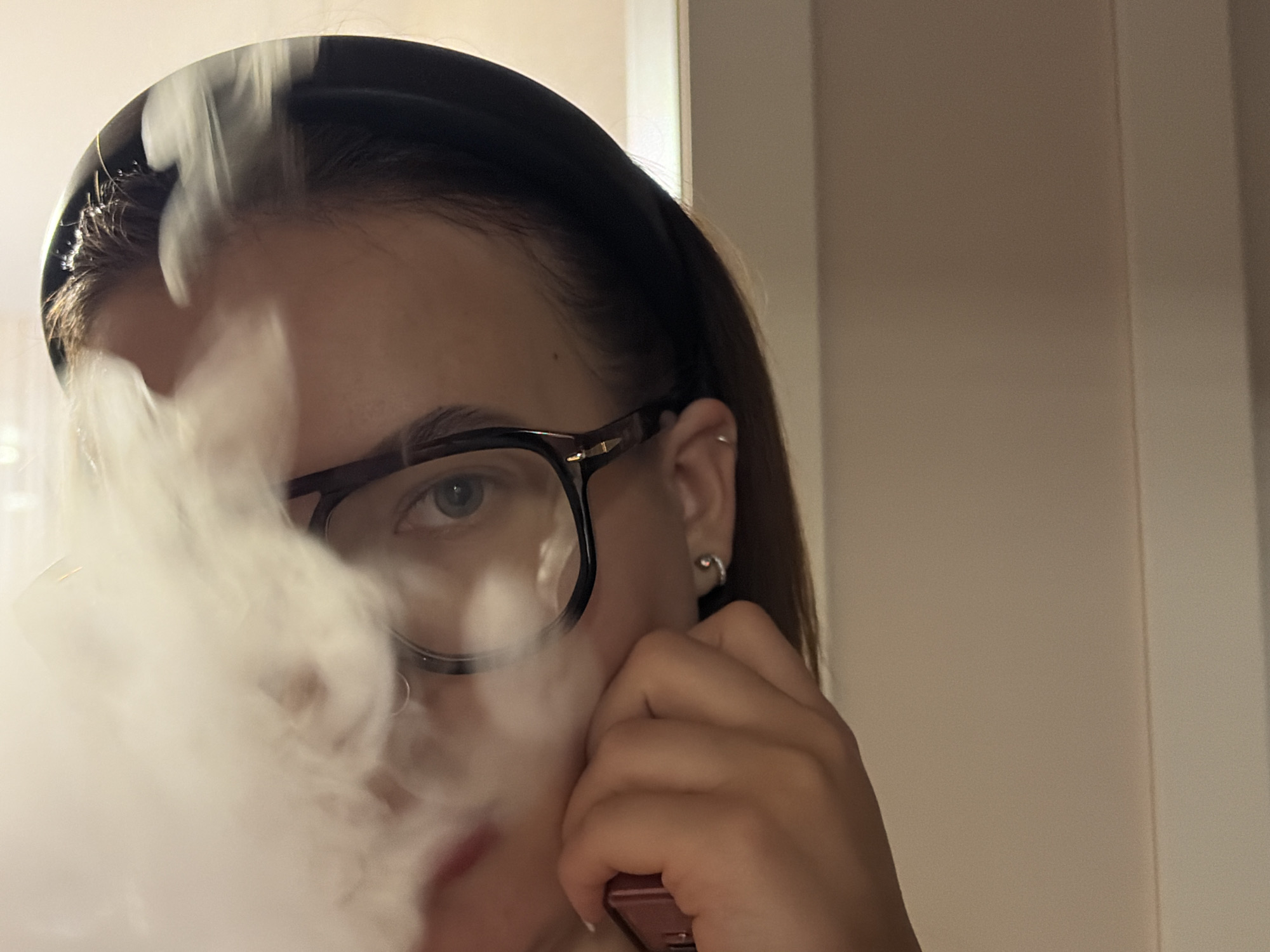 A close-up, slightly angled indoor shot features a young female, partially obscured by a large, swirling plume of white smoke. She wears a dark hairband, pulling back her dark hair, and black-framed glasses. Her left eye, with a light iris, is clearly visible through the glasses, looking directly at the camera. Two small, silver-colored earrings adorn her left ear: a hoop and a smaller stud or ring. Her right hand is visible on the lower right, holding a small, dark red or maroon electronic device, positioned near where the smoke appears to be exhaled, strongly suggesting she is vaping. The background consists of a plain, light-colored wall with a vertical lighter strip on the right, possibly a door frame or room corner. The lighting is somewhat dim, creating an intimate atmosphere. There is no visible text in the image. The scene takes place in Krasnodar, Russia.