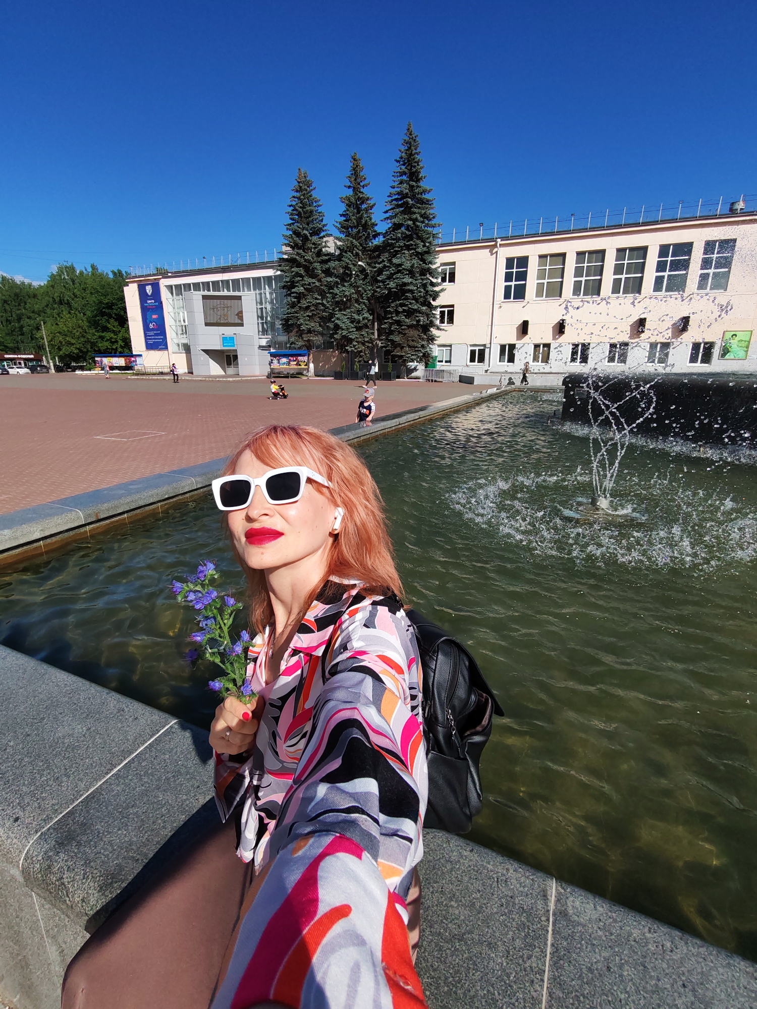 A woman with pink hair takes a selfie in front of a fountain in Chaikovsky, Russia. She is wearing large white sunglasses, a colorful patterned shirt, and a black backpack. She holds a small bouquet of purple flowers in her right hand. The fountain is in the foreground, with water spraying upwards. In the background, there is a light-colored building with many windows, trees, and a clear blue sky. People can be seen in the distance walking in a plaza area. The sun is shining brightly, indicating it is daytime and likely warm weather. The overall mood of the image is bright and cheerful. Visible text includes "60" on a banner on the building.