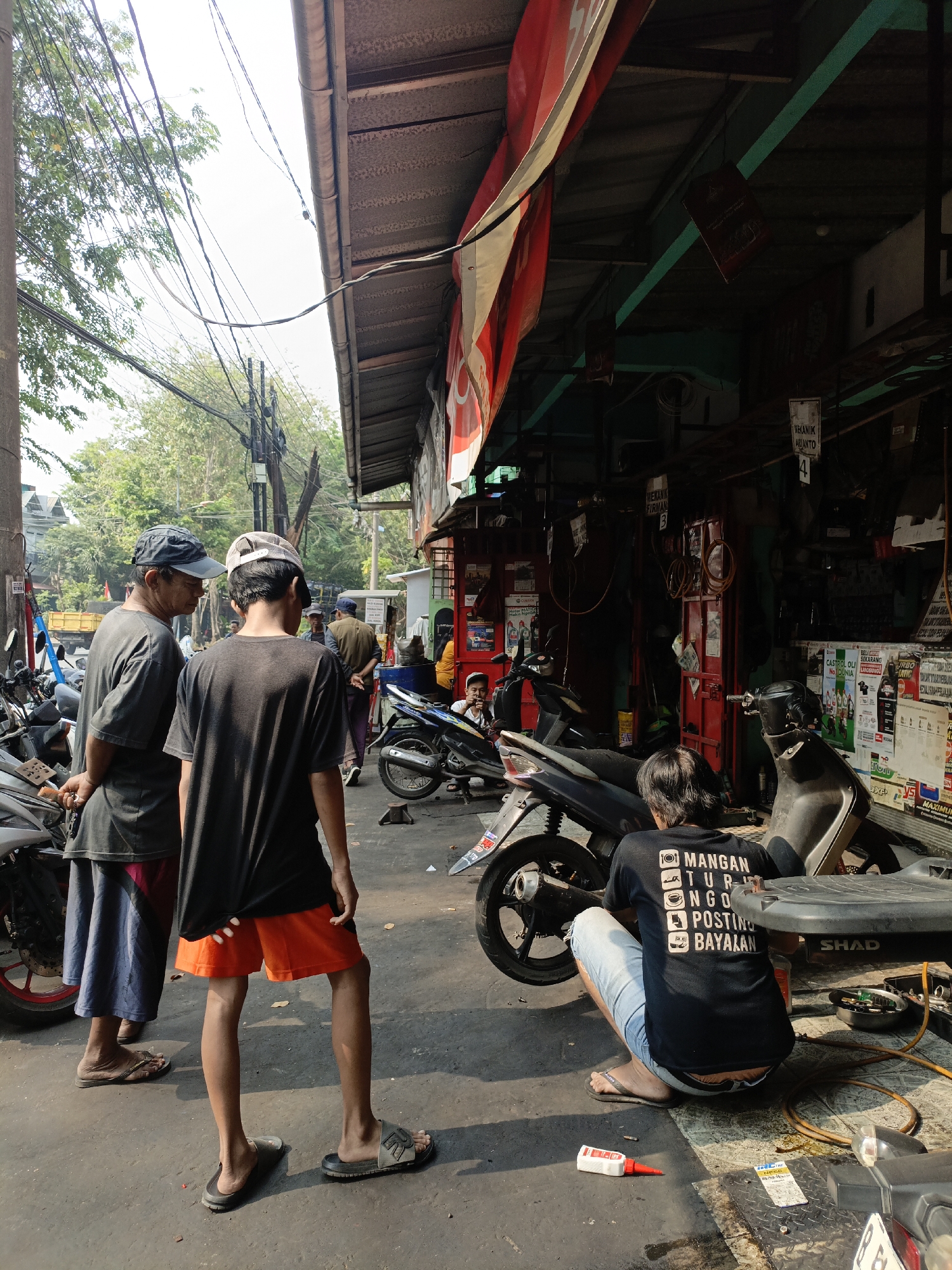 The image depicts an outdoor scene, likely a motorcycle repair shop or a street vendor's stall, in the city of East Jakarta, Indonesia. The foreground features three individuals. Two men are standing, facing away from the camera and appearing to converse. One is older, wearing a dark t-shirt, a dark baseball cap, and a blue sarong-like garment. The younger man next to him wears a black t-shirt and orange shorts. To the right, a third person, also facing away from the camera, is kneeling beside a black motorcycle. This individual wears a black t-shirt with white text and a pair of light blue jeans. The t-shirt text appears to be a list of activities or statements.
The environment is characterized by a narrow street or alleyway with a covered overhang providing shade. The overhang appears to be made of corrugated metal or similar material and is supported by a structure with green beams visible. The walls behind the overhang are lined with posters and advertisements. Several motorcycles are parked around the area, some of them appearing to be under repair. Electrical wires are strung overhead, a common sight in urban Indonesian settings. The ground is paved, and there are various tools and supplies scattered around, including a tube of red and white adhesive glue. Sunlight casts distinct shadows, indicating a bright day. In the background, more buildings and trees are visible, suggesting a densely populated urban area. The overall impression is of a busy, informal commercial space.