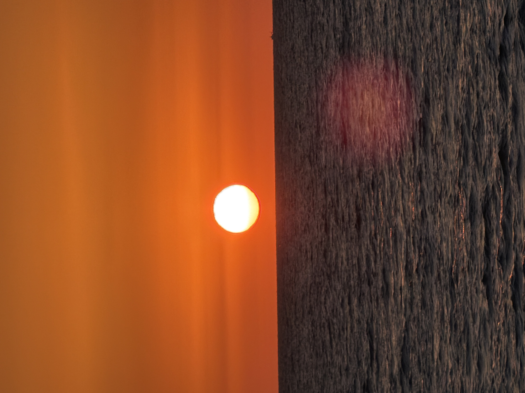 This image captures a sunset over the Mediterranean Sea from the coast of Tel-Aviv, Israel. The sky occupies the upper two-thirds of the vertical frame and is dominated by a clear, gradient expanse of warm hues, transitioning from a medium orange at the top to a vibrant, intense red-orange closer to the horizon. Horizontal atmospheric bands of slightly varying intensity are discernible across the sky. The sun, a brilliant, uniformly bright white circular disk, is positioned slightly above the horizon, to the left of the vertical center. Its intense light creates a subtle glow around its perimeter. The horizon line is sharply defined and perfectly flat, separating the vivid sky from the darker sea below. The lower third of the image depicts the sea, which is a dark slate grey or deep blue-grey, with numerous small ripples and waves texturing its surface. Minimal highlights of reddish-orange sunlight are visible on the wave crests, particularly in the foreground. A distinct, diffused reddish-pink lens flare or internal reflection is visible on the water's surface in the lower right quadrant. In the far distance, on the horizon towards the right, a small, dark silhouette of a ship or boat is present. No people are visible in the scene.