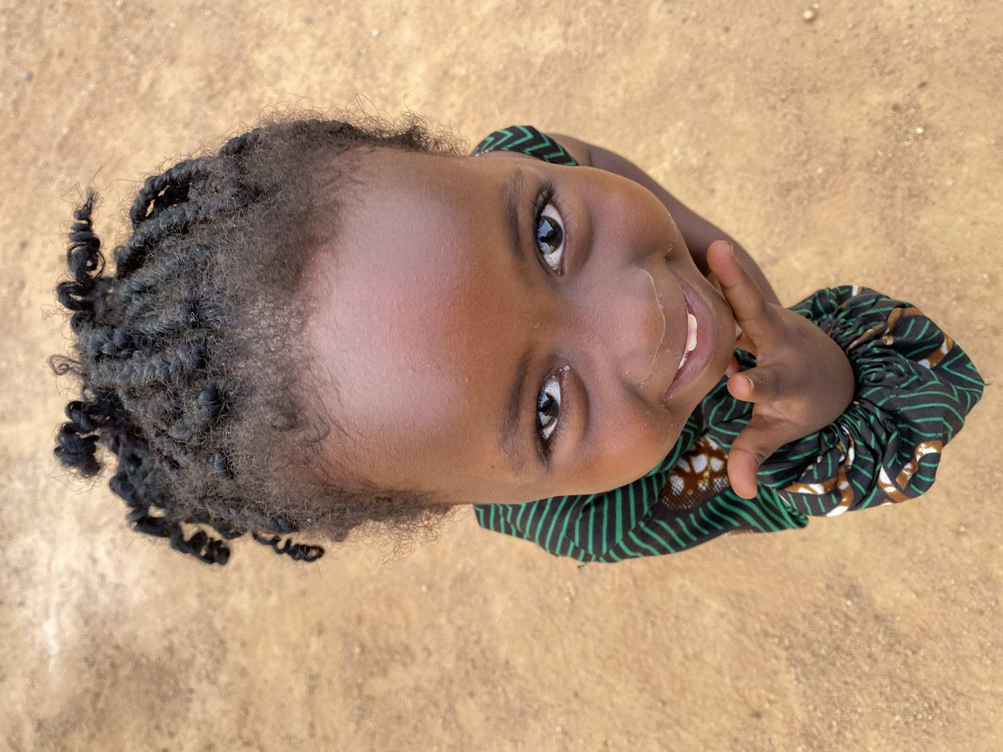 A young, dark-skinned child with small, dark eyes looks upwards, smiling and touching their face with their right hand. The child's hair is dark, tightly curled, and gathered in short twists. They are wearing a garment with a dark green and black striped pattern, with some brown and white accents. The background is a flat, light brown surface, seemingly outdoors. The scene is from Kuru Dungus, Nigeria.