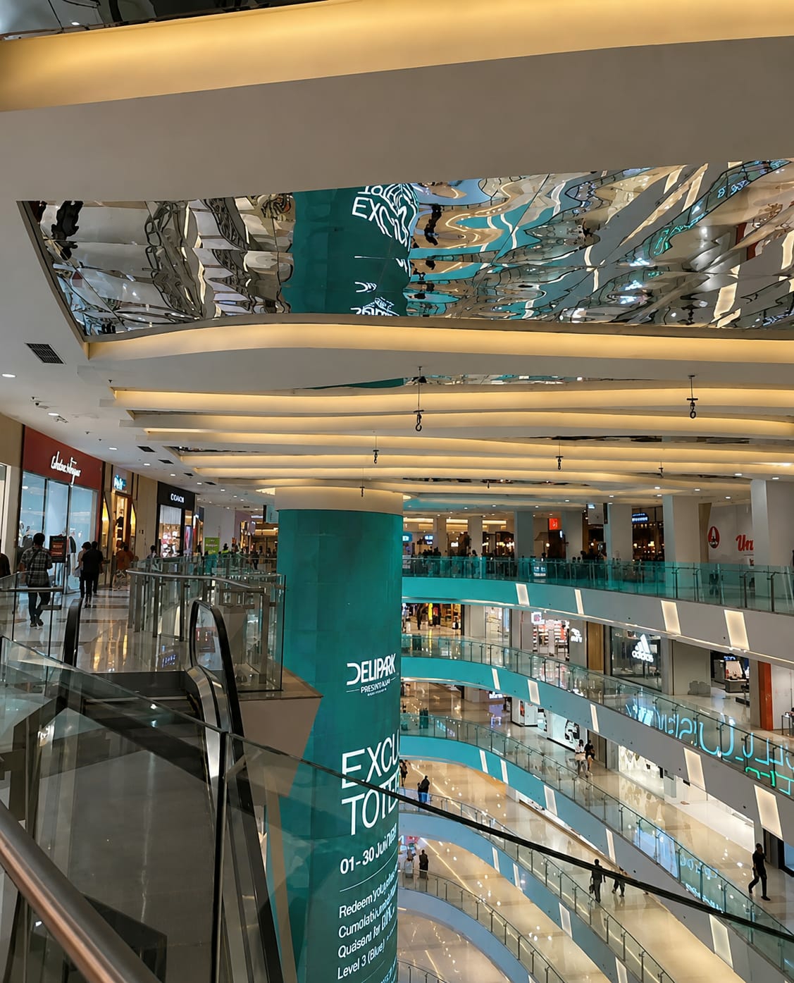 This image depicts the interior of the Delipark Mall in the city of Medan, Indonesia. The scene captures a multi-level shopping mall with a modern architectural design, characterized by curved walkways, glass railings, and sleek lighting. A large cylindrical pillar in the foreground displays promotional text, including "DELIPARK" and "PRESENTS." The mall's interior is filled with various retail stores and escalators, with several shoppers visible on different levels, engaging in typical mall activities such as walking and browsing. The ceiling features a reflective surface with integrated lighting, creating a sense of spaciousness and reflecting the mall's vibrant atmosphere. The overall impression is that of a busy and upscale commercial environment.
