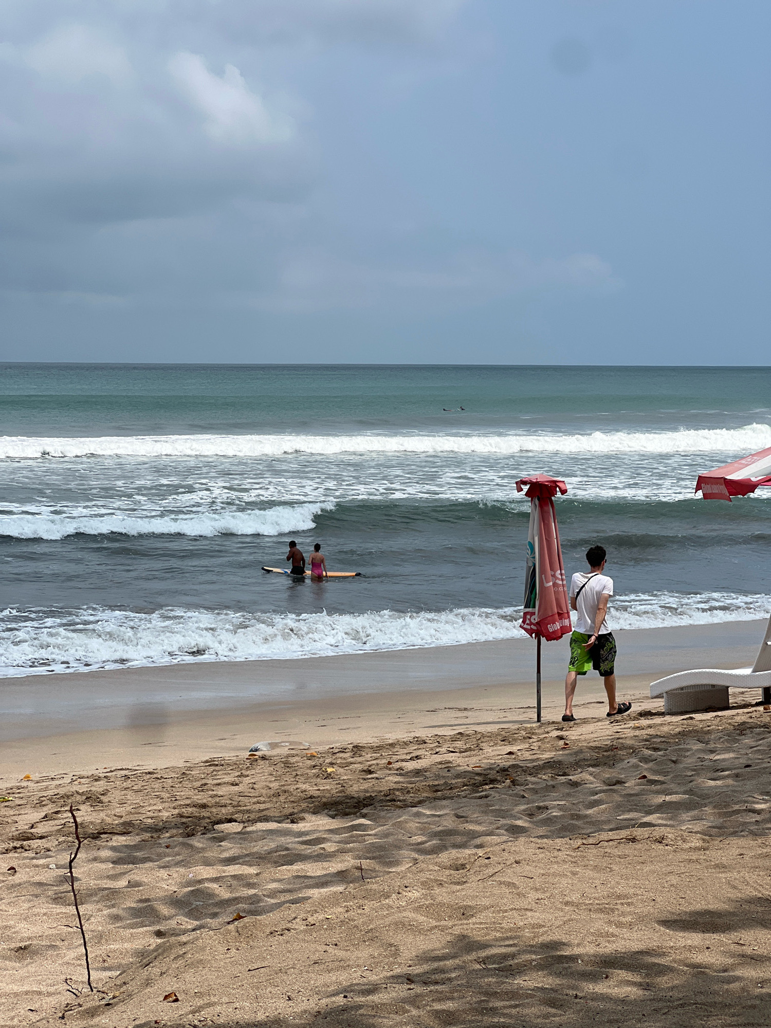 This image depicts a beach scene in Kuta, Indonesia, under a cloudy sky. In the foreground, a sandy beach with scattered debris and dry branches is visible. A person wearing a white t-shirt and green patterned shorts stands facing the ocean, looking out at the water. Next to them is a red and white striped umbrella and a white lounge chair. In the middle ground, two individuals are seated on a surfboard in the shallow water. Further out, the ocean exhibits white waves crashing onto the shore. The horizon is defined by the sea meeting the overcast sky. Two small, dark shapes, possibly surfers or swimmers, are faintly visible in the distance on the water.