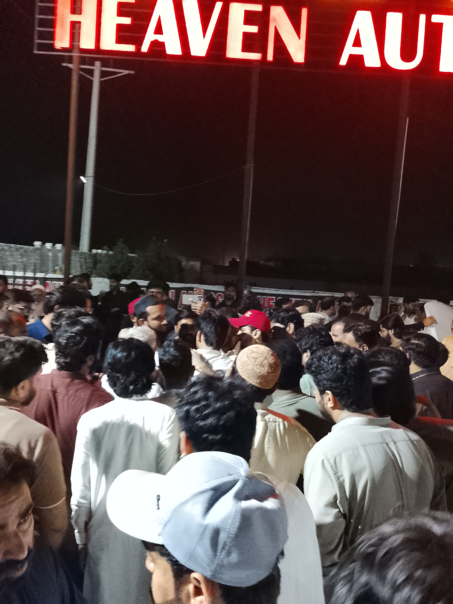 The image captures a dense gathering of men outdoors at night in what appears to be Al Falah Modern City, Pakistan. The scene is illuminated primarily by a large, red neon sign overhead that clearly displays the text "HEAVEN AUTO," suggesting the location is an automotive business or dealership.
The foreground and midground are filled with a large crowd of individuals, mostly men, many of whom are seen from behind or in profile. They are dressed in a variety of clothing, including traditional kurtas and modern attire, with some wearing baseball caps or traditional head coverings. The faces of only a few individuals are partially visible. The crowd appears to be stationary, possibly observing an event or waiting in line, with their attention generally directed towards different points, indicating a collective presence rather than a focused activity like a protest. One person in the mid-background appears to be holding a smartphone, possibly recording or taking pictures.
Utility poles are visible supporting the large sign and extending into the dark background. The sky is black, confirming the nighttime setting. Despite the darkness, the illuminated sign and ambient light provide enough visibility for the crowd to be clearly seen. The overall atmosphere suggests a public gathering at an automotive establishment after dark.