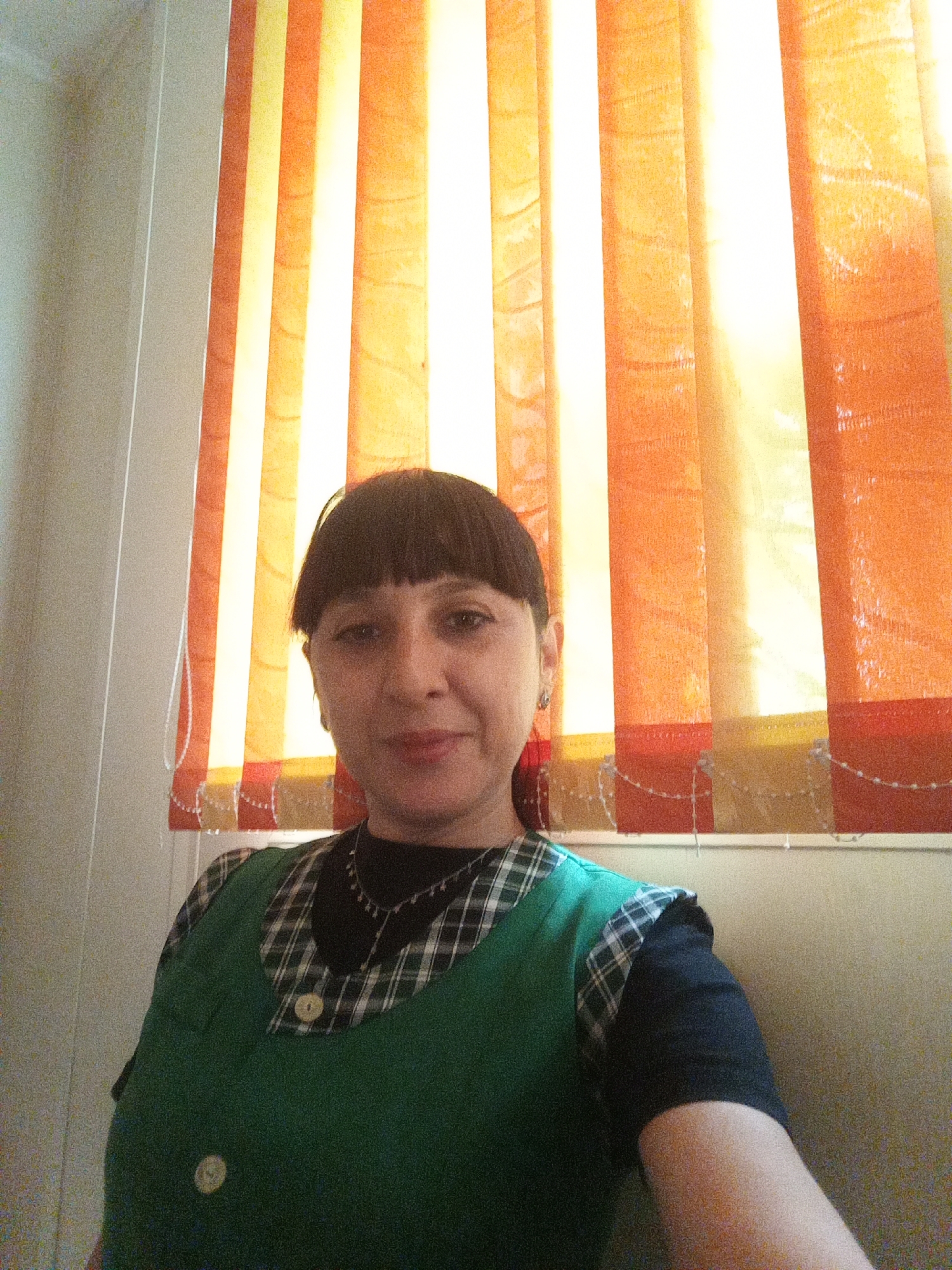 A woman with dark hair and bangs is looking at the camera, wearing a green vest over a black shirt with a plaid collar and a necklace. She is standing in front of vertical blinds that are orange and yellow. The background wall is a light color.