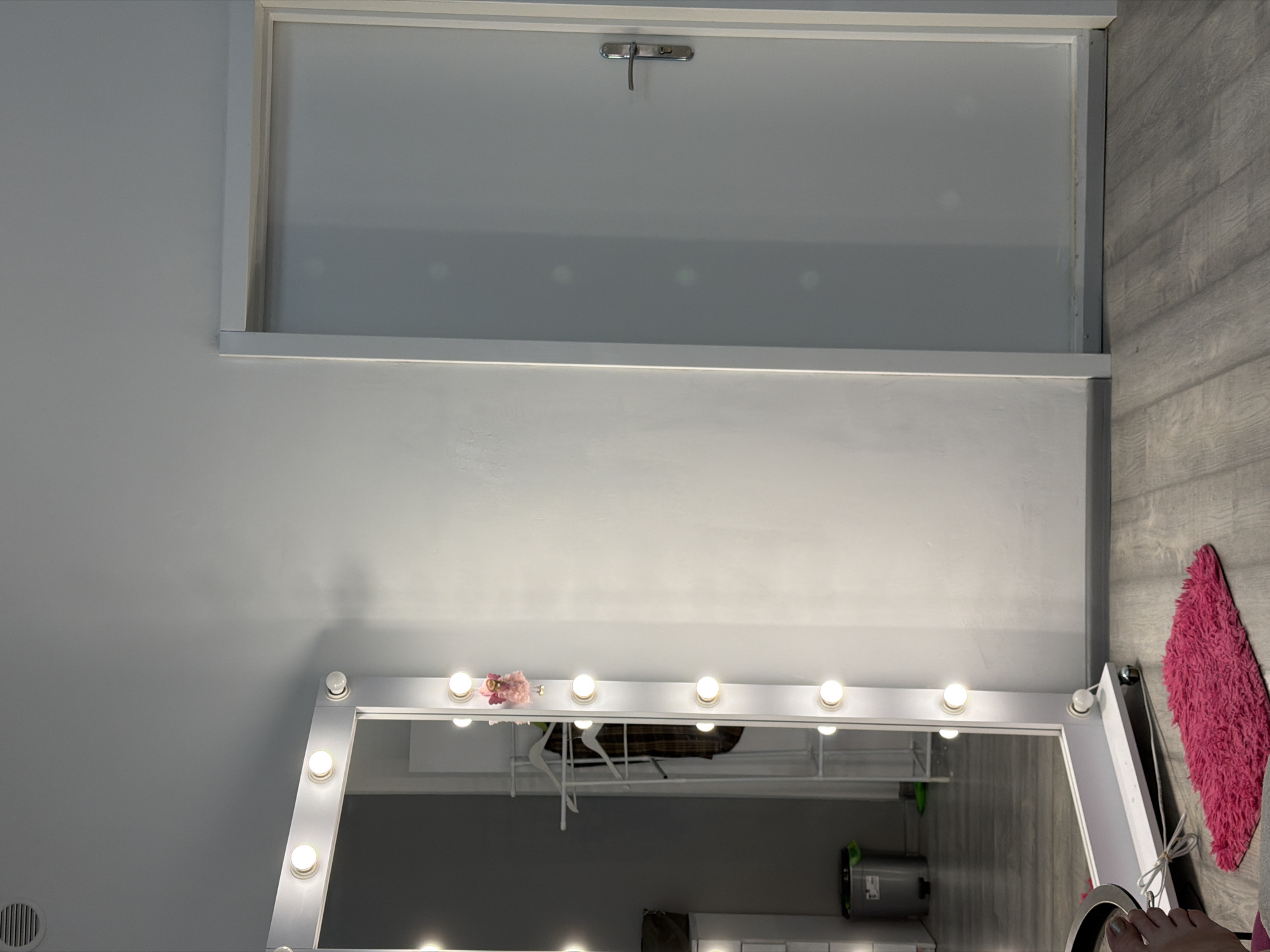 The image depicts an indoor scene, likely a dressing room or a bedroom, with a prominent lighted vanity mirror. The mirror is large and framed in white, with multiple light bulbs around its perimeter. It is positioned at an angle, reflecting a portion of the room, including a clothing rack with garments and a trash can. To the right of the mirror, a white door with a silver handle is visible, suggesting an exit or entrance to another space. The floor is made of grey laminate flooring. A small, bright pink rug is placed in front of the mirror. The walls are painted a light, neutral color. The lighting is primarily provided by the bulbs on the mirror, casting a warm glow. The overall atmosphere appears to be organized and functional, likely set up for personal grooming or preparation.
