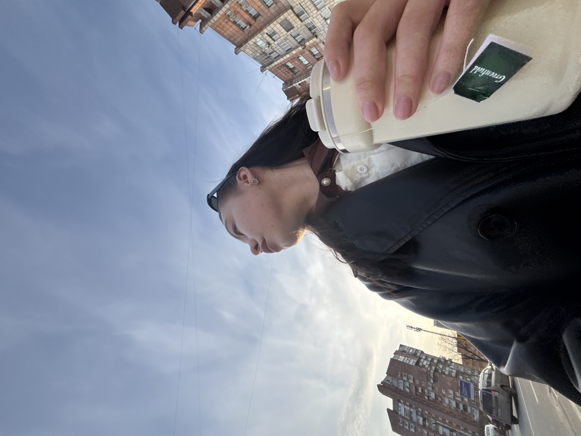 A medium, eye-level shot, tilted to the right, shows a woman holding a beverage container outdoors. The woman, with dark hair pulled back, wears a black leather jacket over a white shirt with brown detailing. A "Greenfield" tea tag is visible on the beverage container. The background includes a cloudy sky, city buildings, and cars on the street. The location is identified as Perm, Russia based on a sign visible on a building behind.