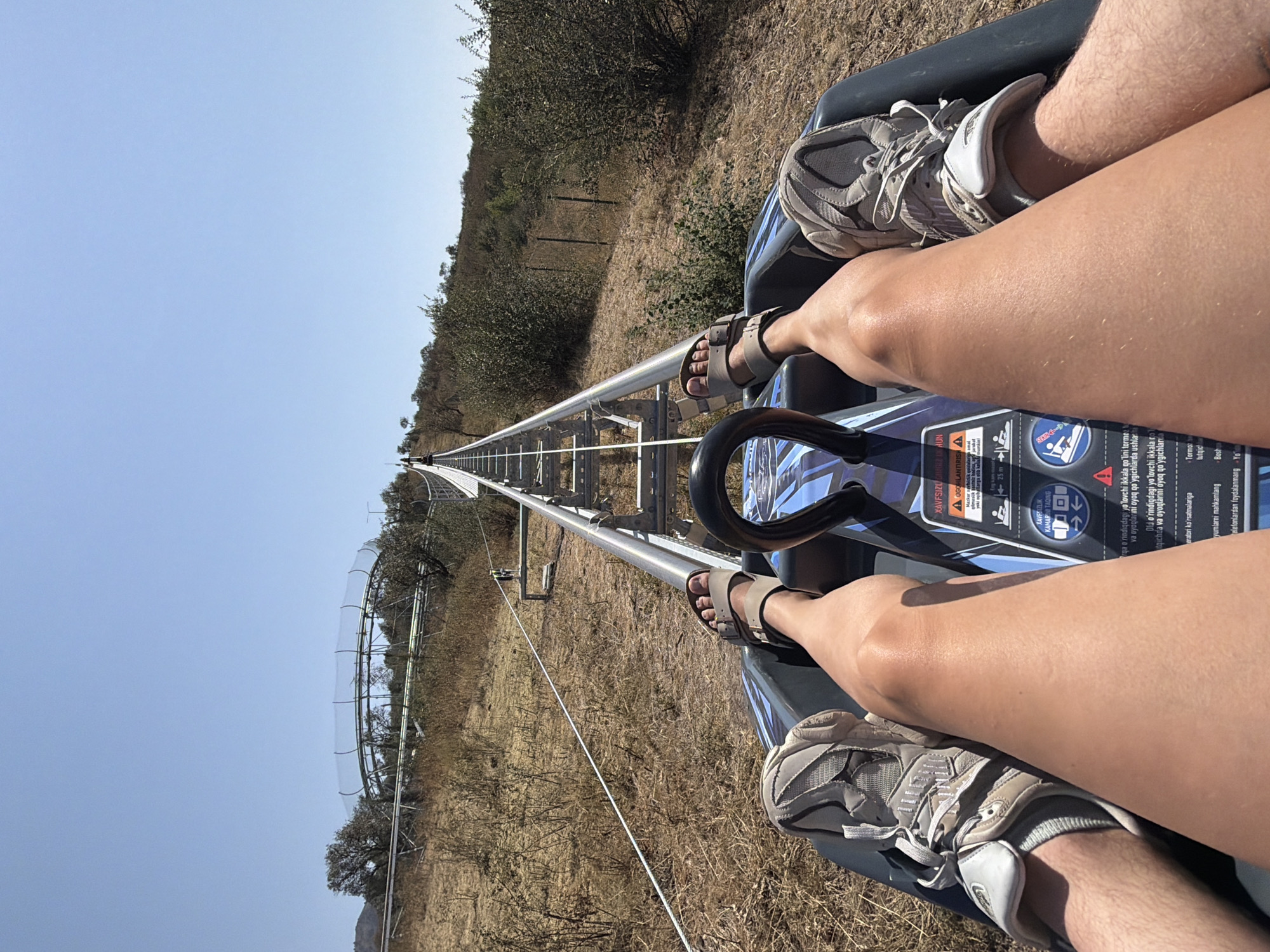 The image captures a first-person perspective of someone riding a bobsled or alpine coaster in the mountainous terrain surrounding Charvak, Uzbekistan. The rider's legs, clad in athletic shoes and sandals, are prominently visible in the foreground, positioned within the bobsled. The bobsled itself is dark gray and features a blue and black graphic design, with a black steering mechanism visible. The bobsled is situated on a metal track that ascends a steep, dry, and grassy hillside dotted with sparse vegetation and trees. In the background, the track continues upwards and curves to the left, with a clear blue sky overhead. The overall environment appears arid and mountainous, characteristic of the region around Charvak.