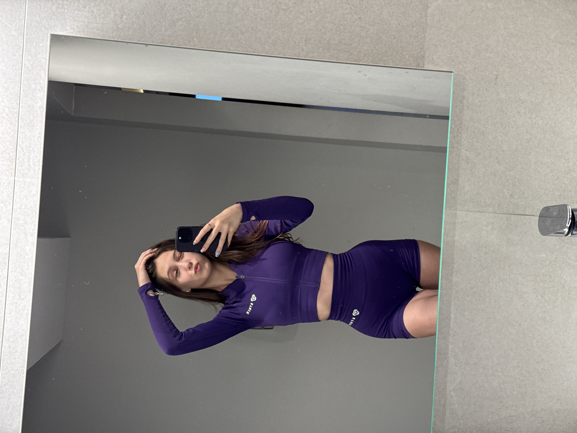 A woman is reflected in a mirror. The woman is wearing a dark purple long-sleeved crop top and matching shorts. She is holding a smartphone in her left hand, taking a picture of herself in the mirror. The background consists of a gray tiled wall and a small chrome fixture. The location is likely a bathroom. The image was taken indoors, but there is not enough information to determine a more specific location within the city of Sofia, Bulgaria.