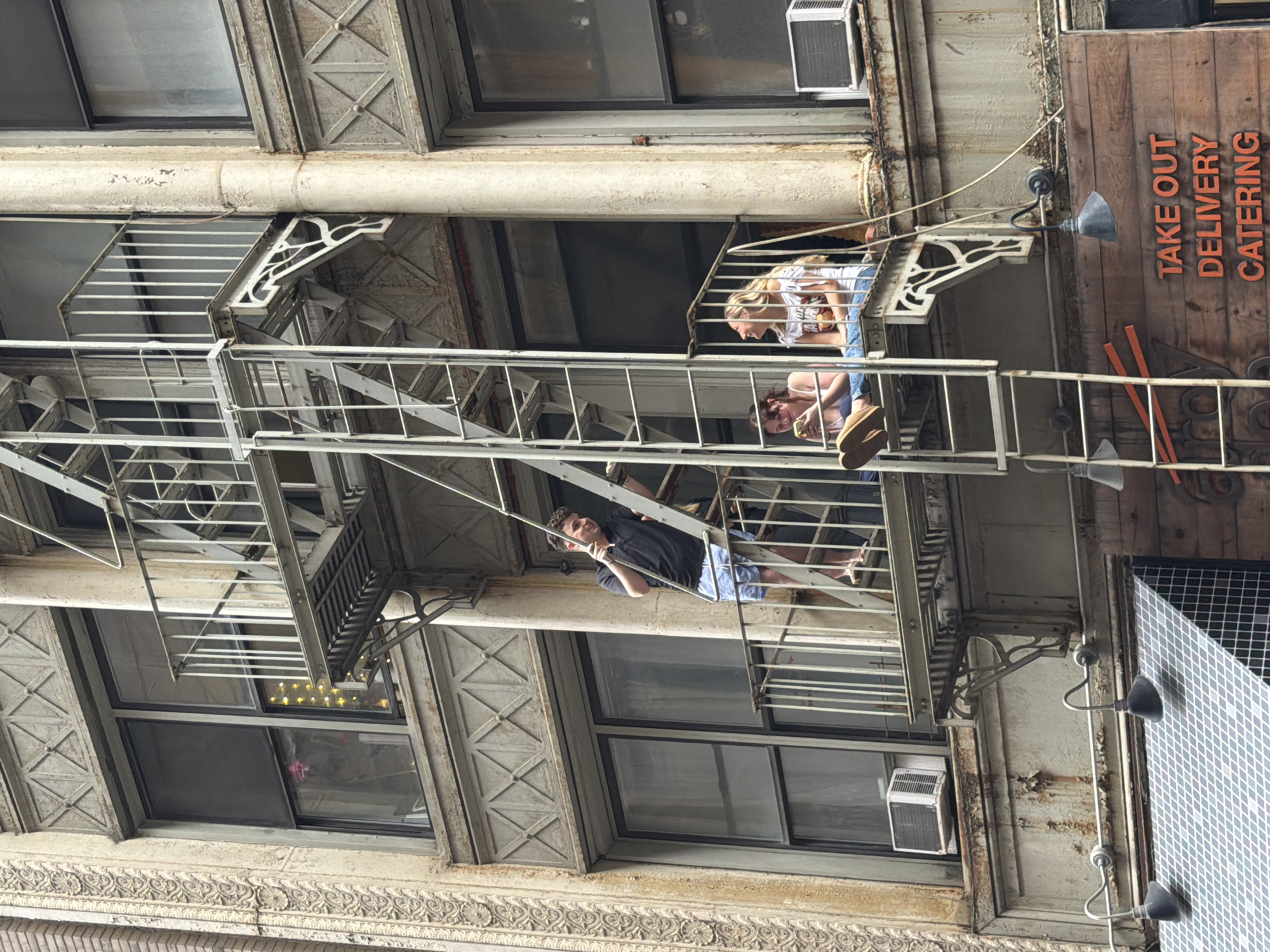 This media file depicts an urban scene featuring a fire escape on an apartment building. The perspective is from below, looking up at several levels of the building. On one of the fire escape landings, a young woman with blonde hair is leaning on the railing, looking downwards. She is wearing a white t-shirt and blue jeans. Below her, on a lower level of the fire escape, another person with dark hair is visible, leaning forward and looking downwards as well, holding something in their hands. On the lowest visible fire escape, a young man with curly dark hair is smiling and looking upwards, leaning on the railing with one hand. He is wearing a black t-shirt and blue shorts.
The building itself appears to be made of brick and has multiple windows. Some windows have air conditioning units installed. The fire escape is made of metal and has a weathered appearance. On the right side of the image, a wooden sign is visible with orange text that reads "TAKE OUT DELIVERY CATERING." The lighting suggests it is daytime, possibly with overcast conditions. The overall scene conveys a sense of casual urban life, with people interacting and enjoying the outdoors from their balconies or fire escapes. The building's architectural details, such as the decorative cornices and window frames, suggest an older structure, potentially in a historic part of New York City.