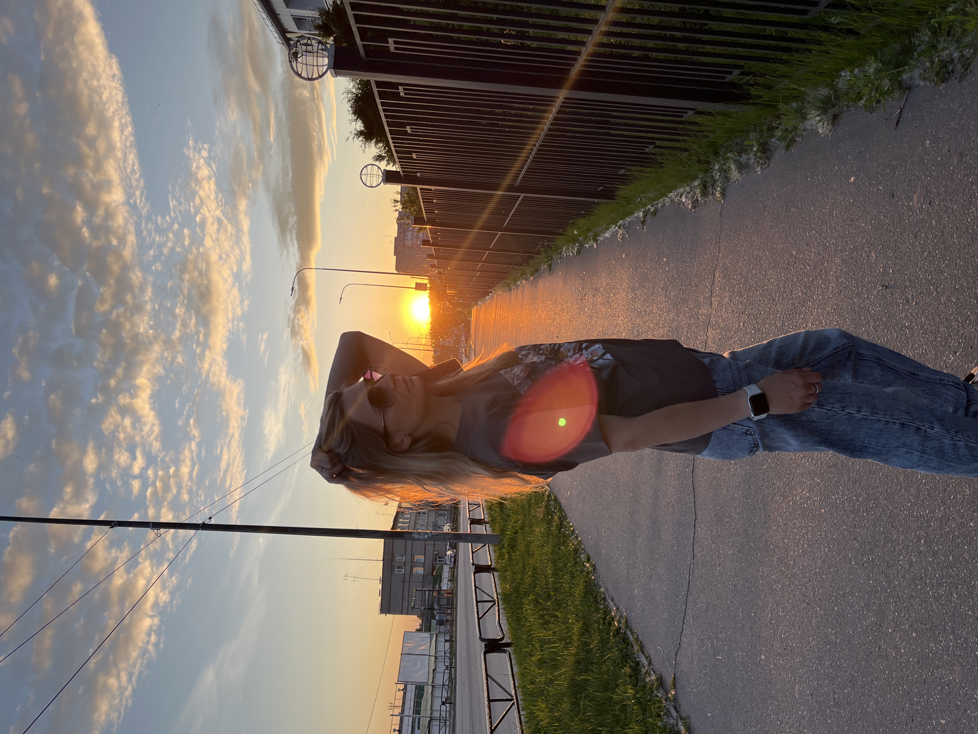A woman with long blonde hair, wearing sunglasses, a dark t-shirt, and blue jeans, stands in profile on an asphalt sidewalk. Her right hand is raised, fingers intertwined in her hair. She wears a silver smartwatch on her left wrist and a ring on her left ring finger. The setting sun, a bright orange orb, is visible directly behind her to the right, casting a strong orange glow on her hair and shoulders, and a sun flare is visible in the lower-left quadrant of her torso.
To her left, there is a narrow strip of green grass and a low black metal barrier separating the sidewalk from a multi-lane road. Across the road, several low-rise, utilitarian-style buildings are visible, along with utility poles and wires, and distant vehicles. To her right, a tall black metal fence runs parallel to the sidewalk, also separated by a strip of grass. Globe-shaped decorative elements top some of the fence posts. Streetlights are visible along both sides of the street. The sky above is a gradient of blue, fading to orange and yellow near the horizon, with scattered, textured altocumulus clouds. The ground shows cracks in the asphalt and some white, fluffy debris (possibly poplar fluff) along the edges. The scene depicts an evening or early morning view, illuminated by the low sun.
Based on the provided information, the location is in the city of Chaikovsky, Russia. The architecture of the buildings, typical of post-Soviet urban development, aligns with this geographical context.