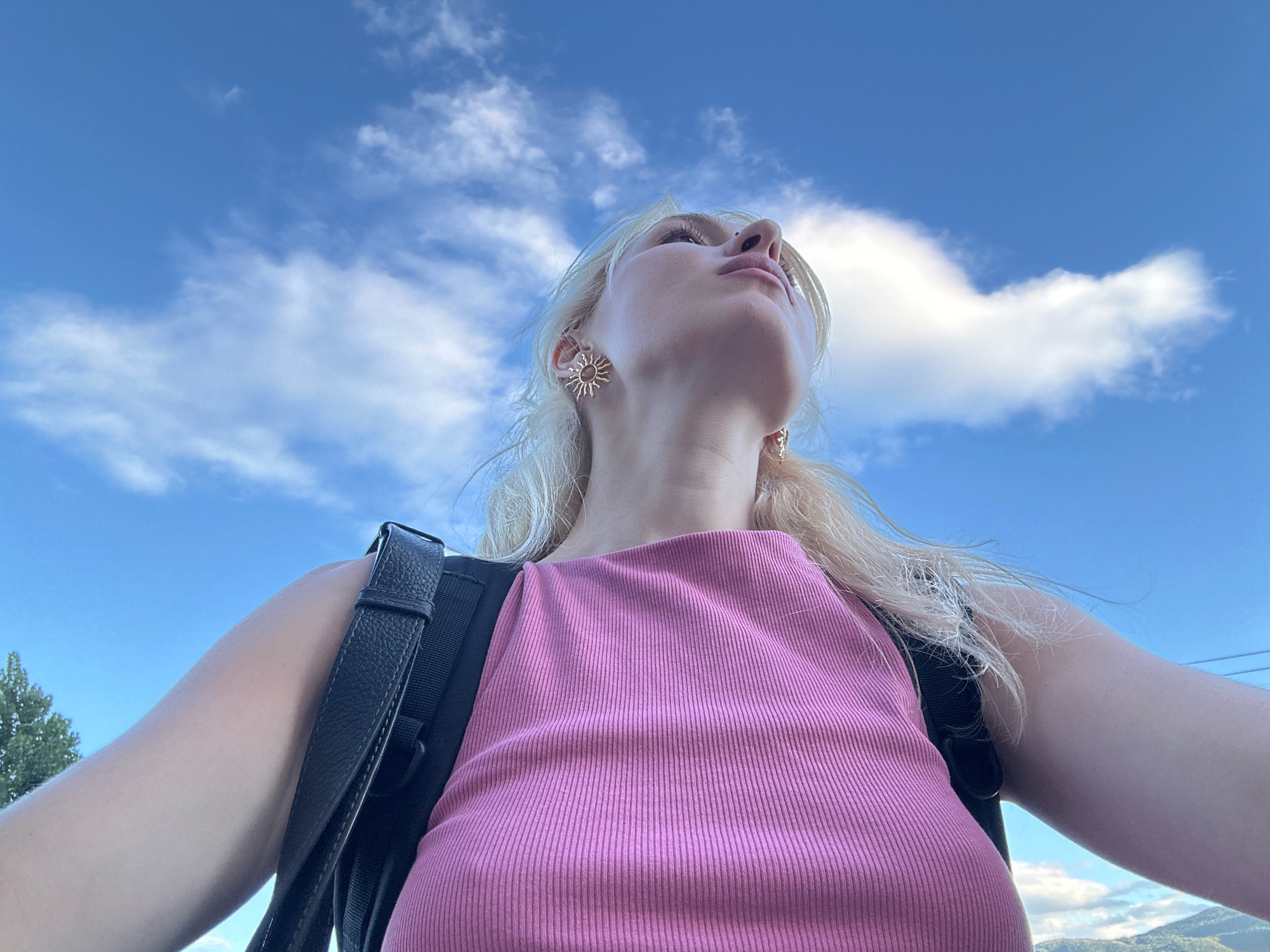 An outdoor, low-angle, selfie-style photograph features a young Caucasian woman with blonde hair and fair skin. She is looking upwards with her head tilted back. She wears a pink, ribbed tank top and a black backpack with visible straps. She has gold, sun-shaped earrings. The background is dominated by a bright blue sky with scattered white clouds. A sliver of green foliage is visible on the left side of the frame, and a hint of a mountainous landscape with a few power lines can be seen at the bottom right. The lighting suggests it is daytime. There is no visible indication of the location being in Tbilisi, Georgia.