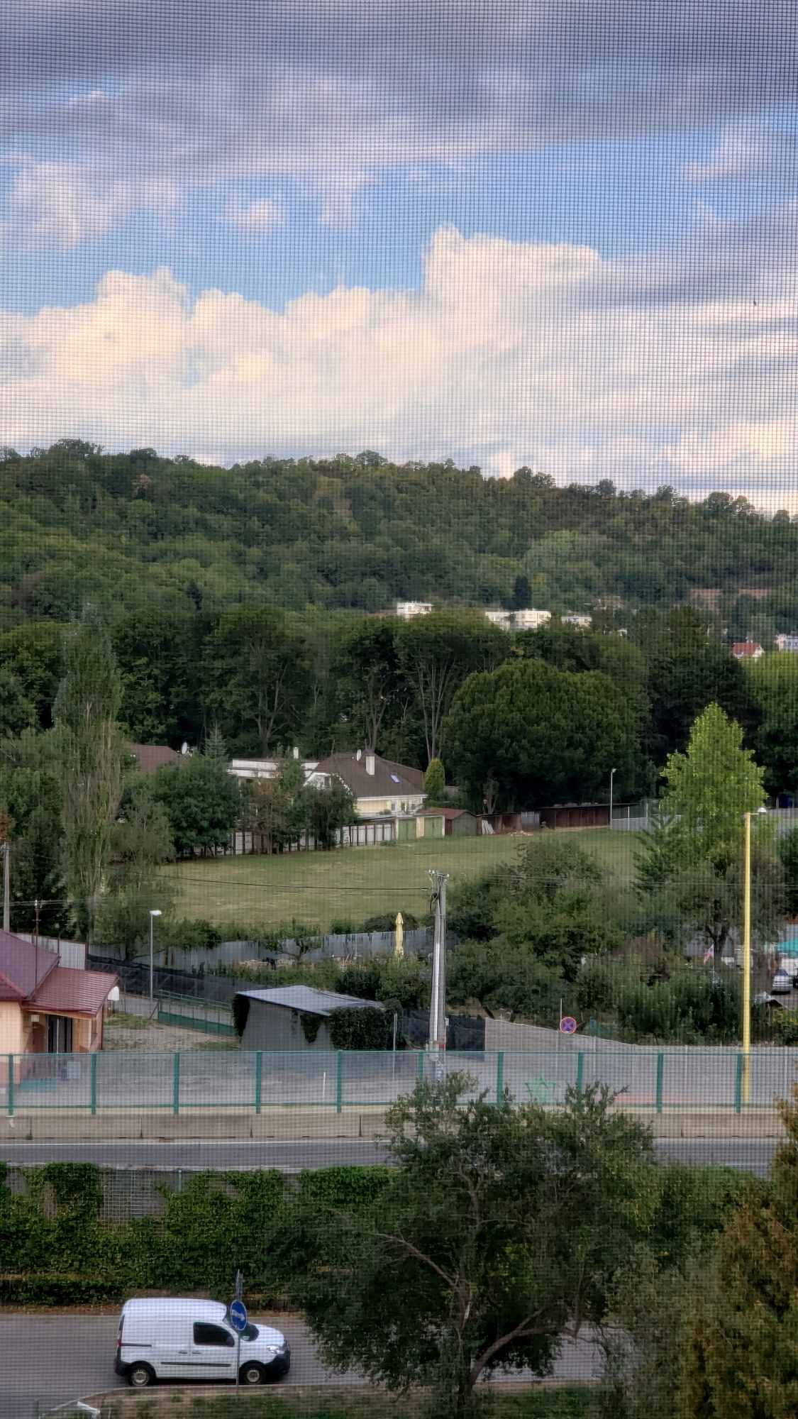 This is a medium-angle, vertical, eye-level shot taken through a window screen, showing a view of a suburban area in Prešov, Slovakia, on an overcast day. In the foreground, a white van is parked on a street. Beyond the street, a green sound barrier separates it from a grassy field with several small sheds. Further back, a residential area with houses and trees is visible, nestled at the base of a densely wooded hillside. The sky above is filled with scattered clouds.