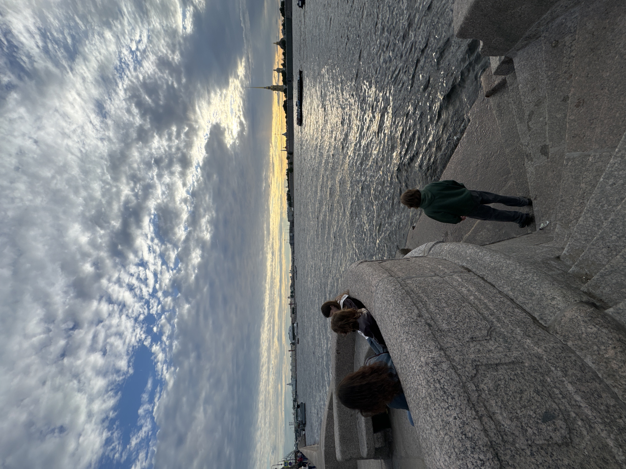 The image depicts a view of the Neva River in Saint Petersburg, Russia, at sunset. In the foreground, a curved granite embankment with a built-in bench overlooks the water. Three individuals, viewed from behind, are seated on the bench. A fourth individual, also viewed from behind, stands on granite steps leading down to the river. The river is choppy with reflected sunlight. In the distance, the Peter and Paul Fortress is visible on the horizon, its spire prominent against a sky filled with dramatic clouds. Several boats are present on the river.