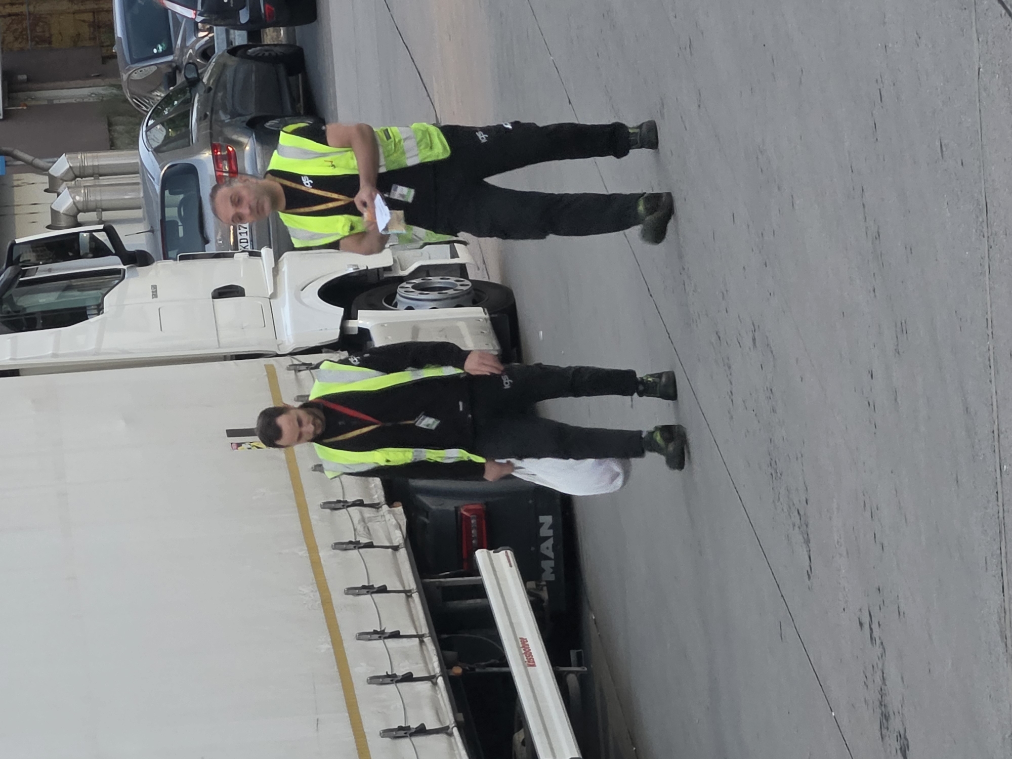 Two men wearing high-visibility vests over black shirts and pants are walking near the side of a white semi-trailer truck. The man on the left is carrying a white plastic bag. The man on the right is holding a small piece of paper and what appears to be a lighter. The truck has "MAN" branding visible on its lower rear panel, and a reflective white strip with "Kässbohrer" printed on it runs along the side. In the background, several cars are parked, including a silver Mercedes-Benz. The scene takes place on a paved surface, likely a street or parking area, in Frankfurt, Germany.