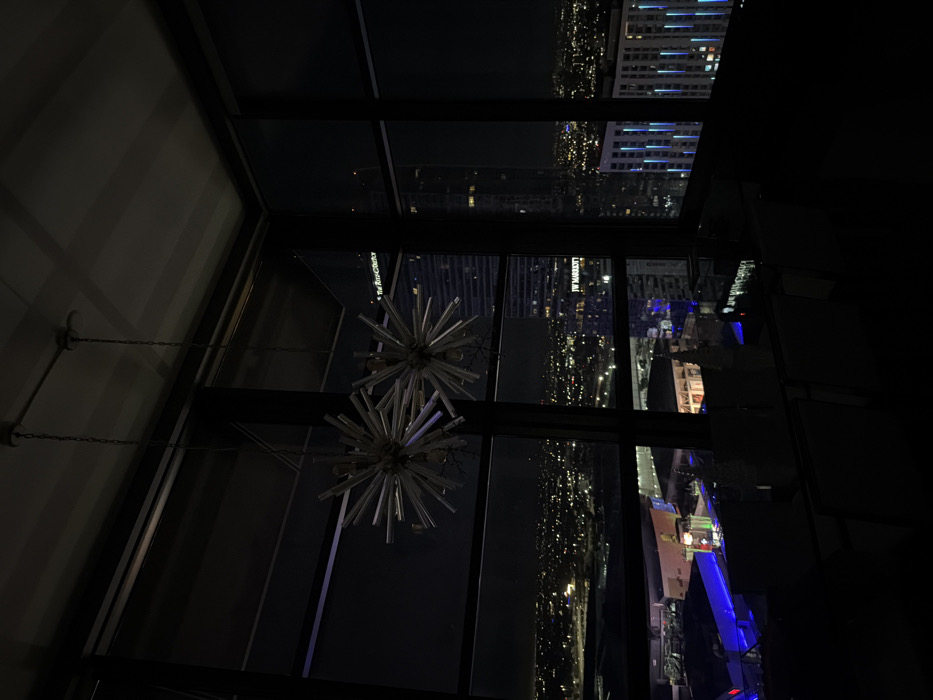 The image presents a nighttime panoramic view of Downtown Los Angeles, United States, observed from an elevated interior space. In the upper-left foreground, two decorative starburst-style chandeliers, composed of numerous radiating cylindrical elements, hang from the ceiling. Large, dark-framed windows dominate the scene, providing an expansive view of the city. A dimly lit table with chairs is partially visible in the lower foreground, featuring several small, white, conical decorative objects. The exterior cityscape is illuminated by countless artificial lights extending into the distance. Prominently visible building signage identifies "JW MARRIOTT" and "The Ritz-Carlton" on a neighboring high-rise, confirming the location as overlooking the L.A. LIVE complex. Another skyscraper exhibits vertical blue accent lighting, and a brightly lit entertainment or commercial area is visible at street level below.