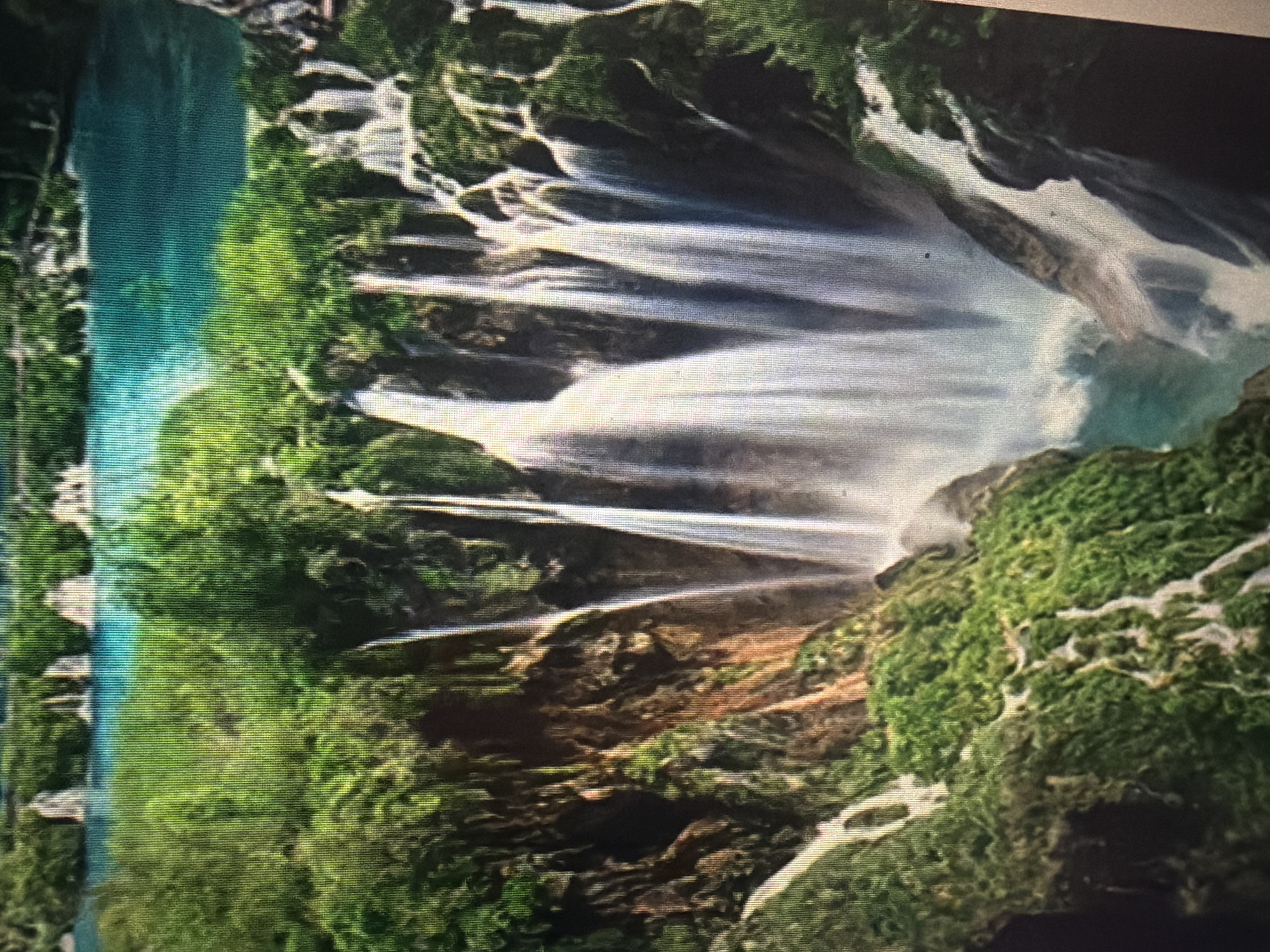 The image displays a picturesque waterfall cascading down lush, green cliffs into a clear turquoise body of water. The scene is a natural landscape with abundant vegetation surrounding the falls. The image does not depict any people, urban elements, or the city of Baan Mae Nam, Thailand. The focus is solely on the natural beauty of the waterfall and its surroundings.