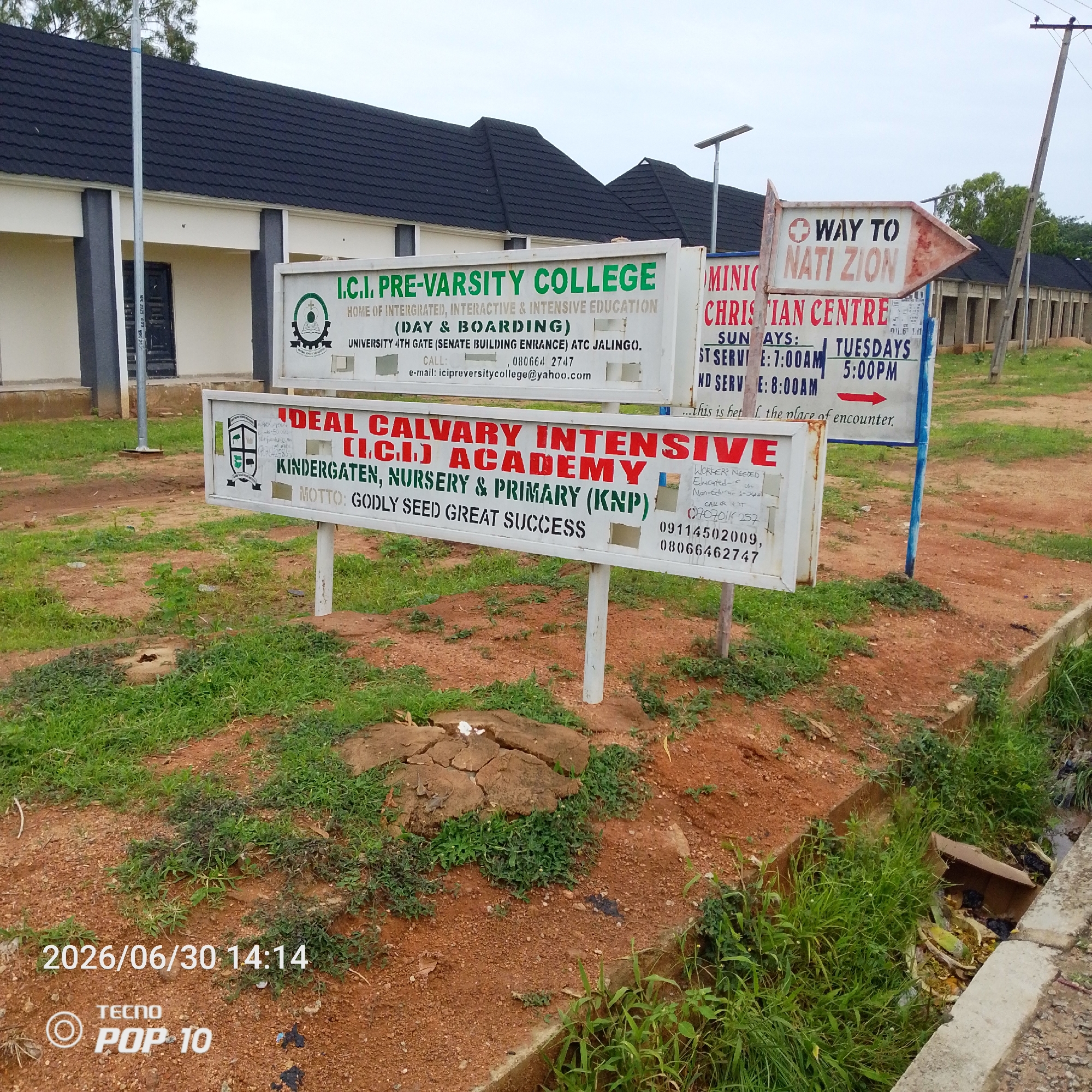 This media file is a photograph taken in Jalingo, Nigeria, showing a street scene with several signs and buildings. The foreground is dominated by a paved ditch filled with grass and some debris, with reddish-brown earth and scattered green vegetation around it. A white sign with a date and time stamp "2026/06/30 14:14" and "TECNO POP 10" is visible in the lower left corner.
In the middle ground, there are three prominent white signs on metal poles. The leftmost sign reads "I.C.I. PRE-VARSITY COLLEGE HOME OF INTERGRATED, INTERACTIVE & INTENSIVE EDUCATION (DAY & BOARDING) UNIVERSITY 4TH GATE (SENATE BUILDING ENRANCE) ATC JALINGO." Below this, contact information including a phone number and email address is listed. The middle sign is for "IDEAL CALVARY INTENSIVE (I.C.I.) ACADEMY KINDERGATEN, NURSERY & PRIMARY (KNP)" with the motto "GODLY SEED GREAT SUCCESS." This sign also includes a list of requirements for workers. The rightmost sign has a directional arrow pointing to the right, indicating "WAY TO NATI ZION" and also displays information about "MINIC CHRISTIAN CENTRE" with service times for Sundays and Tuesdays.
Behind the signs, a building with a dark, textured roof and light-colored walls is visible, featuring dark-colored doors. Further in the background, another structure with a similar roofline and a series of pillars suggests a row of buildings. A utility pole with wires is also present. The overall impression is of an outdoor setting, likely during the daytime, with overcast or cloudy weather. The scene appears to be in a semi-urban or rural area, with educational institutions and a religious center being prominent features. There are no people visible in the photograph.