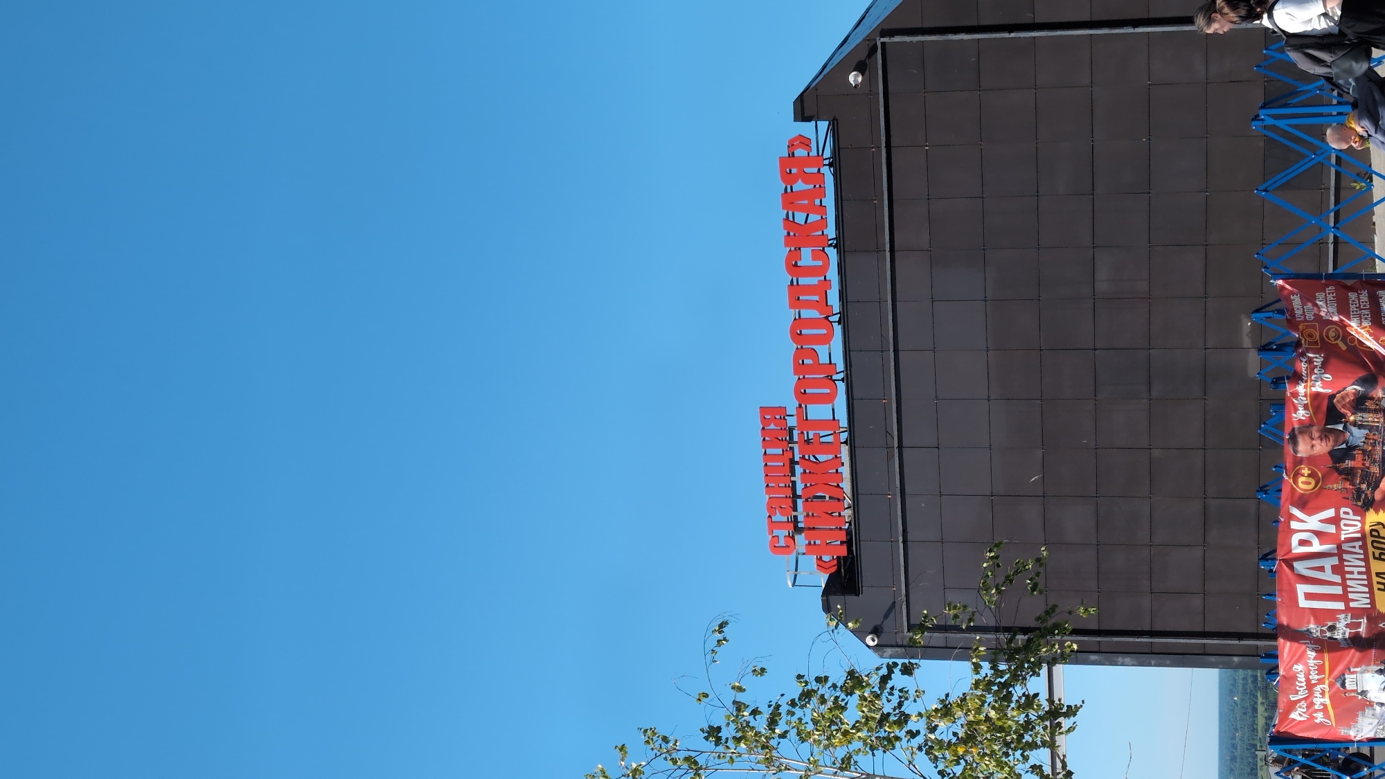 The image shows a clear blue sky above a dark building with red signage. The sign reads "Станция «Нижегородская»" (Station "Nizhegorodskaya"). Below the building, a red banner with various text and images is visible, advertising a "ПАРК МИНИАТЮР НА БОРУ" (Miniature Park on Bor). There are several people visible near the banner, some walking and others with a stroller. To the left, a tree with green foliage is partially in frame. The overall scene suggests an outdoor location in Nizhny Novgorod, Russia, likely near a transportation hub or attraction.