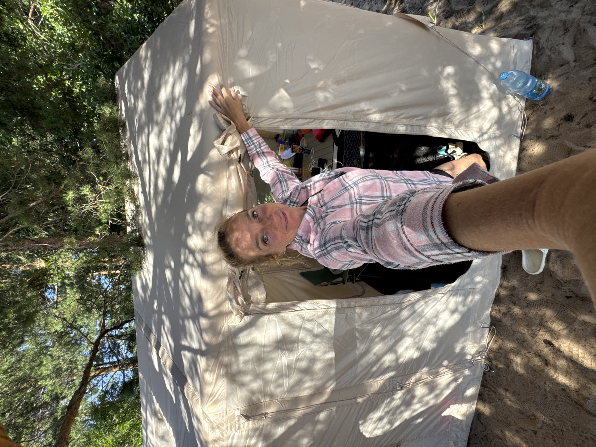 A woman, appearing to be in her late 20s to early 30s, with blonde hair pulled back, is taking a selfie in front of a tan tent. She is wearing a pink and white plaid shirt and large hoop earrings. Her left arm is extended towards the camera, and her right hand is on the tent's zipper. The tent is set up outdoors, possibly in a wooded area, as indicated by the trees and dappled sunlight casting shadows on the tent. Inside the tent, a table is visible with various items, including cups and what appears to be a cooler. A blue plastic water bottle is on the ground to the right of the tent opening. The ground is sandy and has footprints. The overall scene suggests a camping or outdoor recreational setting. The image is taken from a low angle, looking up at the woman and the tent.