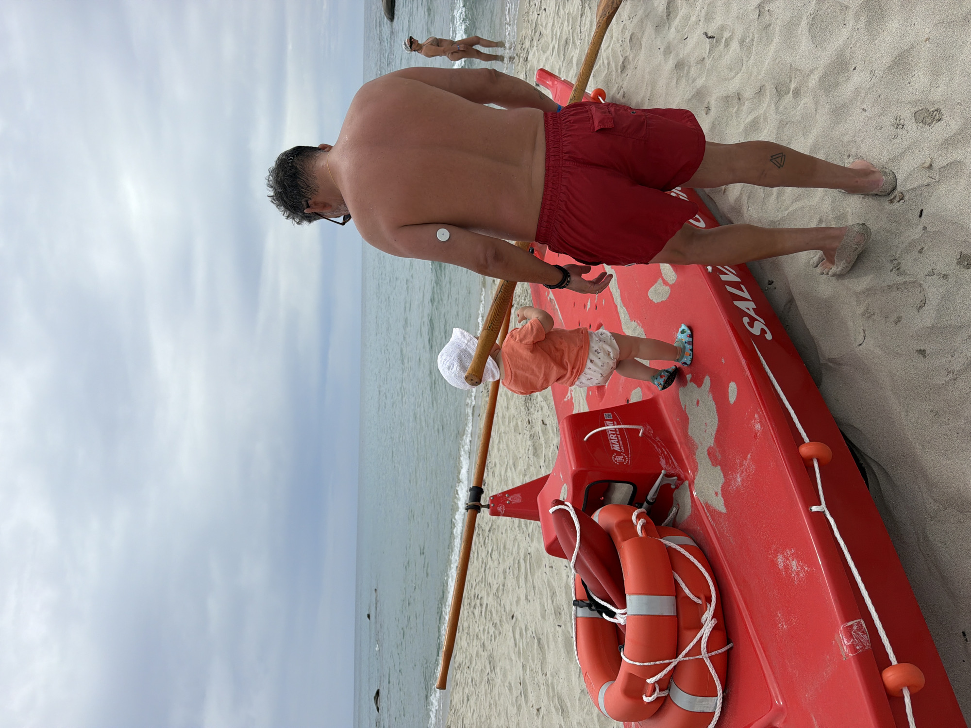 The image depicts a beach scene in Tortolì, Italy, with a red lifeguard boat positioned on the sand near the water's edge. The sky is overcast with gray clouds, and the sea is calm.
In the foreground, a man wearing red swim trunks stands with his back to the camera, facing the sea. He has a small, circular medical device attached to his upper left arm. To his left and slightly in front of him is a young child wearing a white sun hat and an orange t-shirt. The child is looking towards the sea and appears to be holding a wooden oar.
The red lifeguard boat is the central object in the image. It has two orange life preservers stacked on top of it, along with another orange life buoy positioned horizontally. The boat is labeled with the word "SALVE" in white lettering. There are also two wooden oars resting on the boat, extending from the front towards the back.
In the background, a woman is visible standing in the shallow water, facing away from the camera. She is wearing a bikini and a hat. The beach is sandy, with some scattered debris.